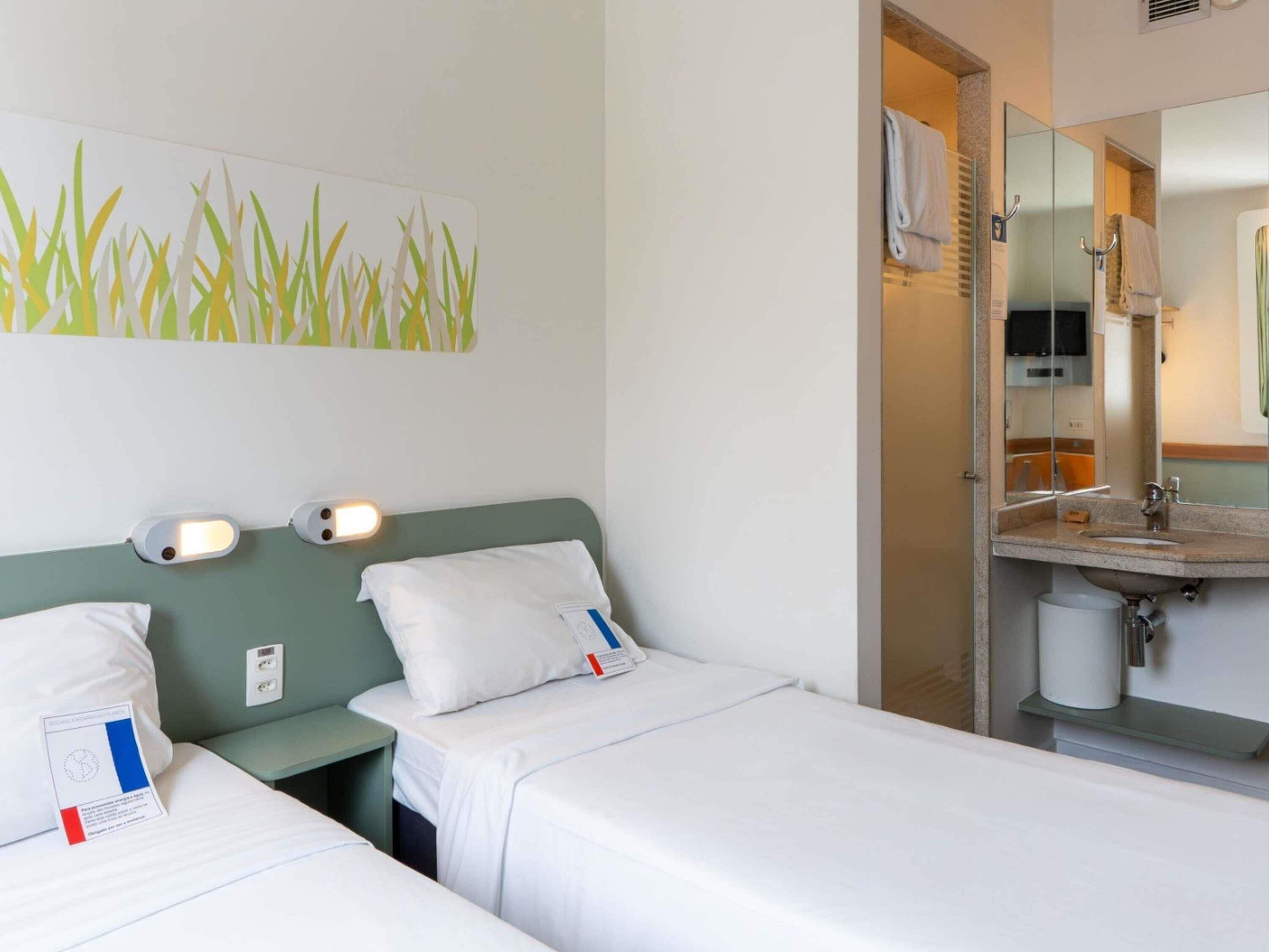 Habitación de hotel con dos camas, un baño con espejo y lavabo, y una pared decorada con una planta.