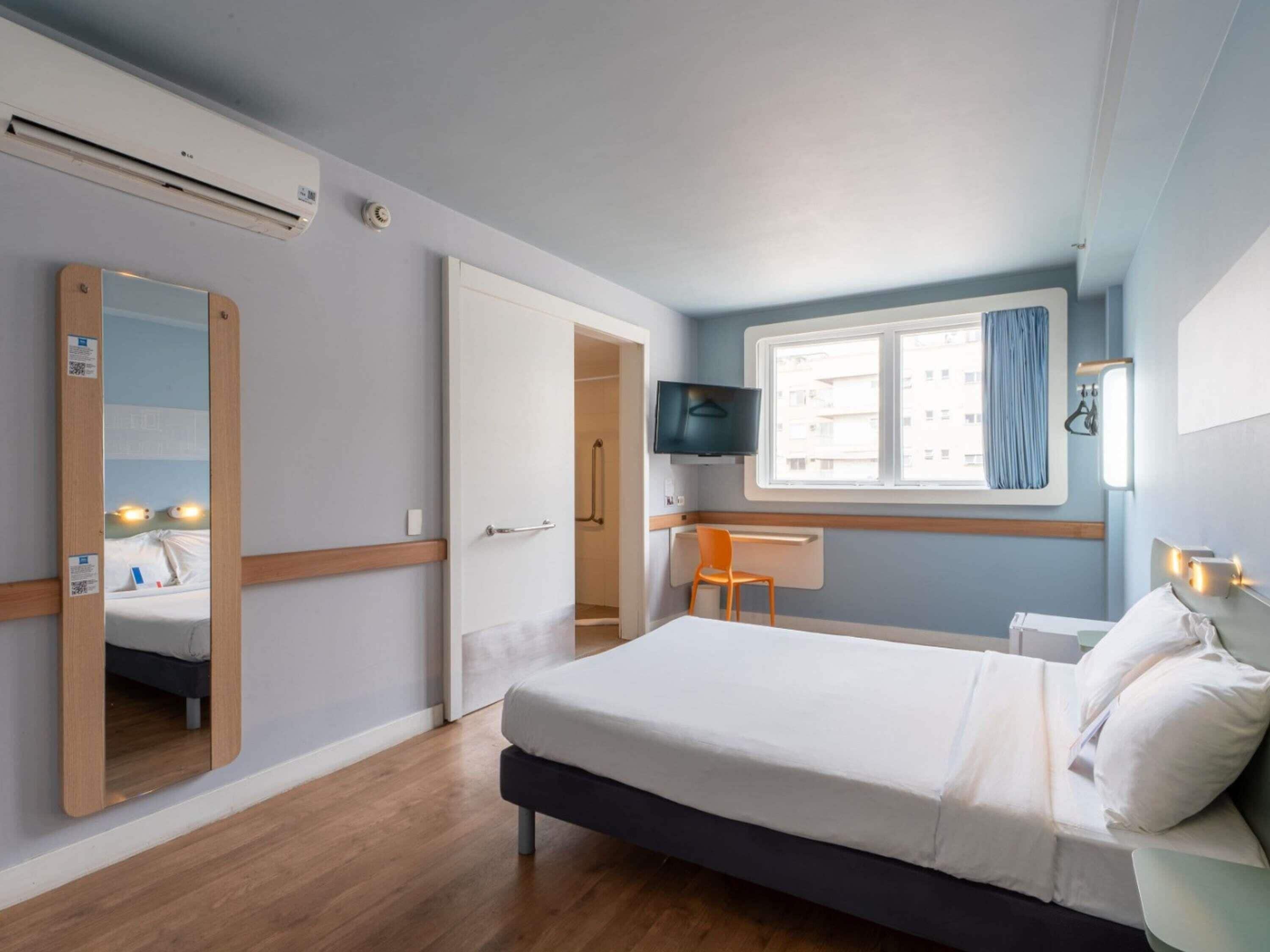 Um quarto com uma cama, televisão, escrivaninha e uma cadeira.