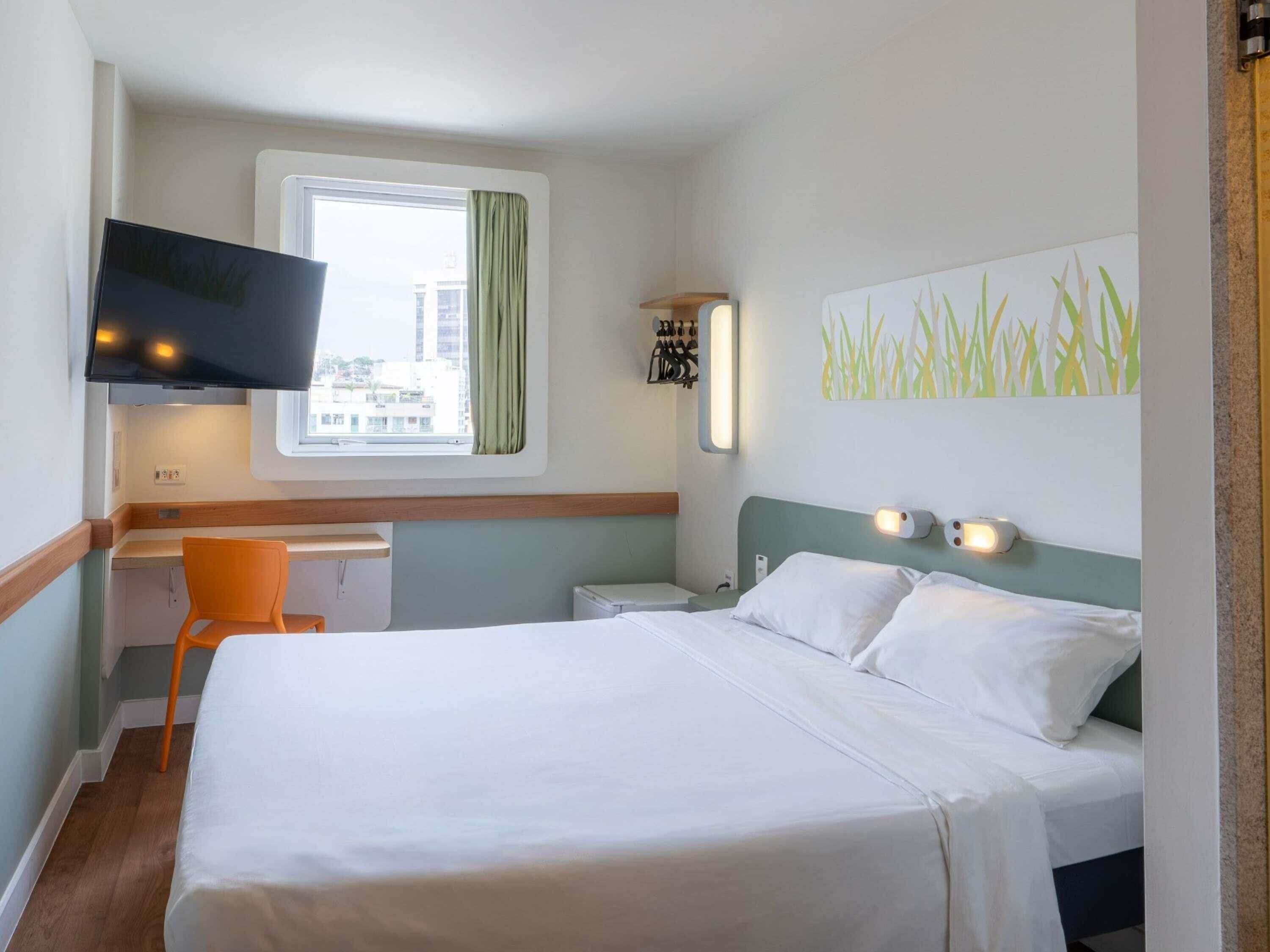 Una habitación de hotel con cama, escritorio, silla, televisor y ventana con cortinas.