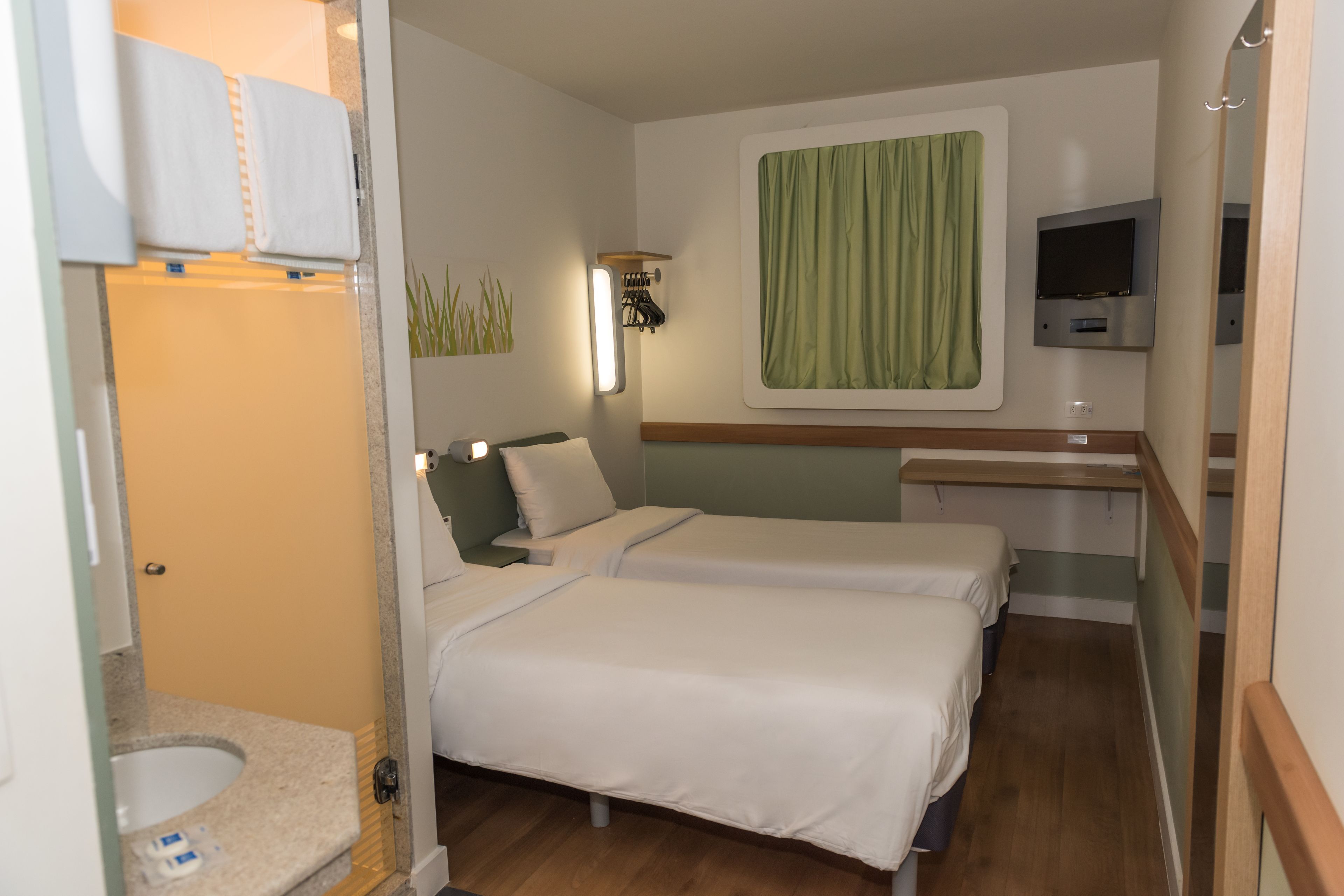 Habitació d'hotel amb dos llits, un bany petit, una televisió i una cortina verda.