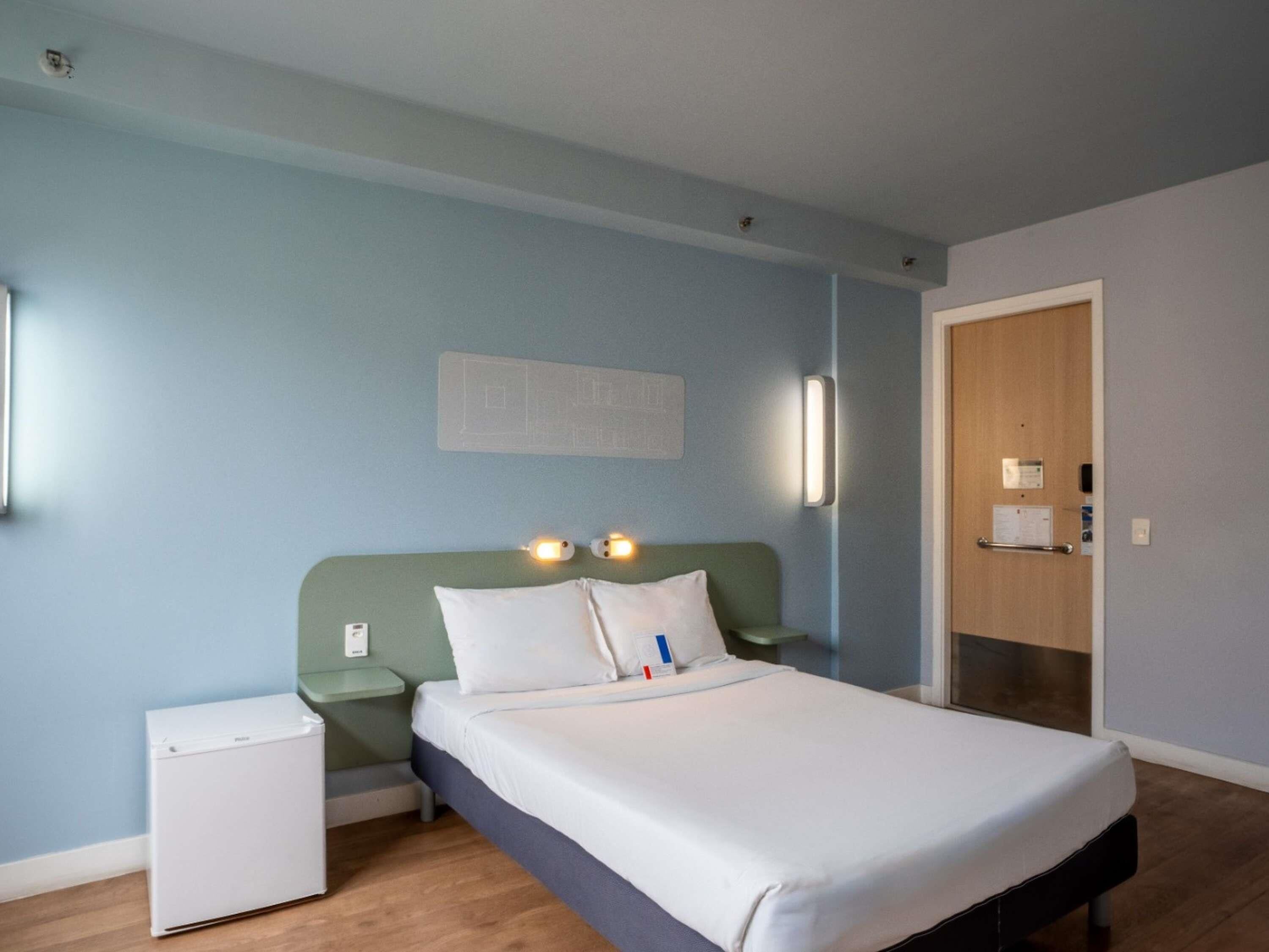 A room with a bed, a nightstand, a small white appliance, and a wooden door.