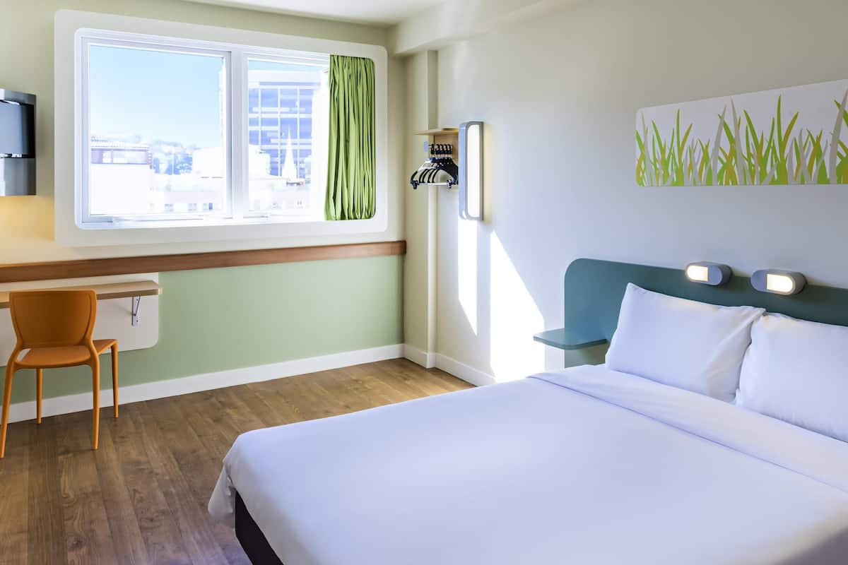 A hotel room with a bed, a desk, a chair, a window with curtains, and a wall-mounted light fixture.