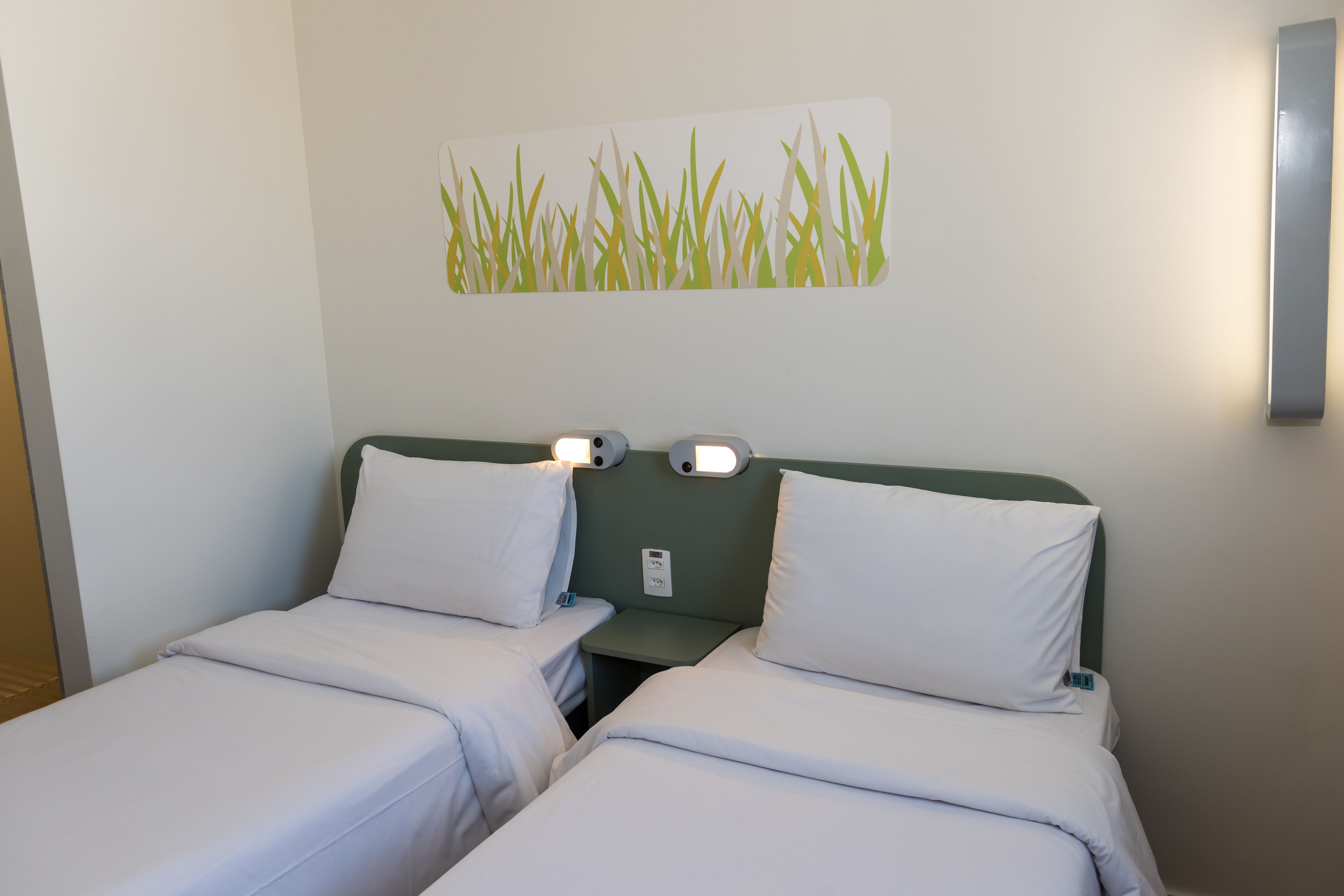 Une chambre d’hôtel avec deux lits simples, chacun doté de draps blancs et d’une tête de lit. Un luminaire est fixé au mur au-dessus des lits, et une petite table se trouve entre les deux.