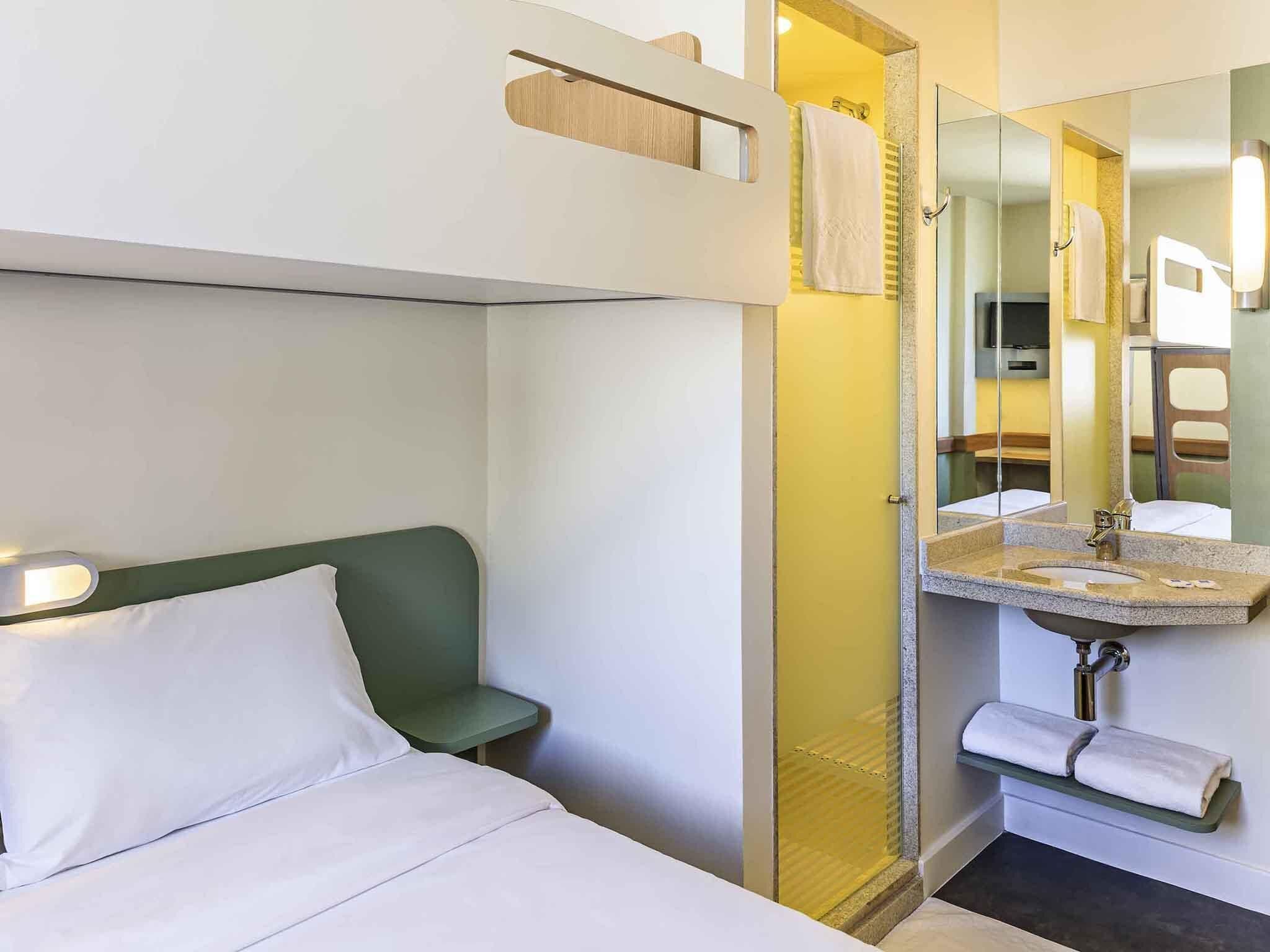 A hotel room with a bed, a green chair, a bathroom with a sink and mirror, and a shower area.