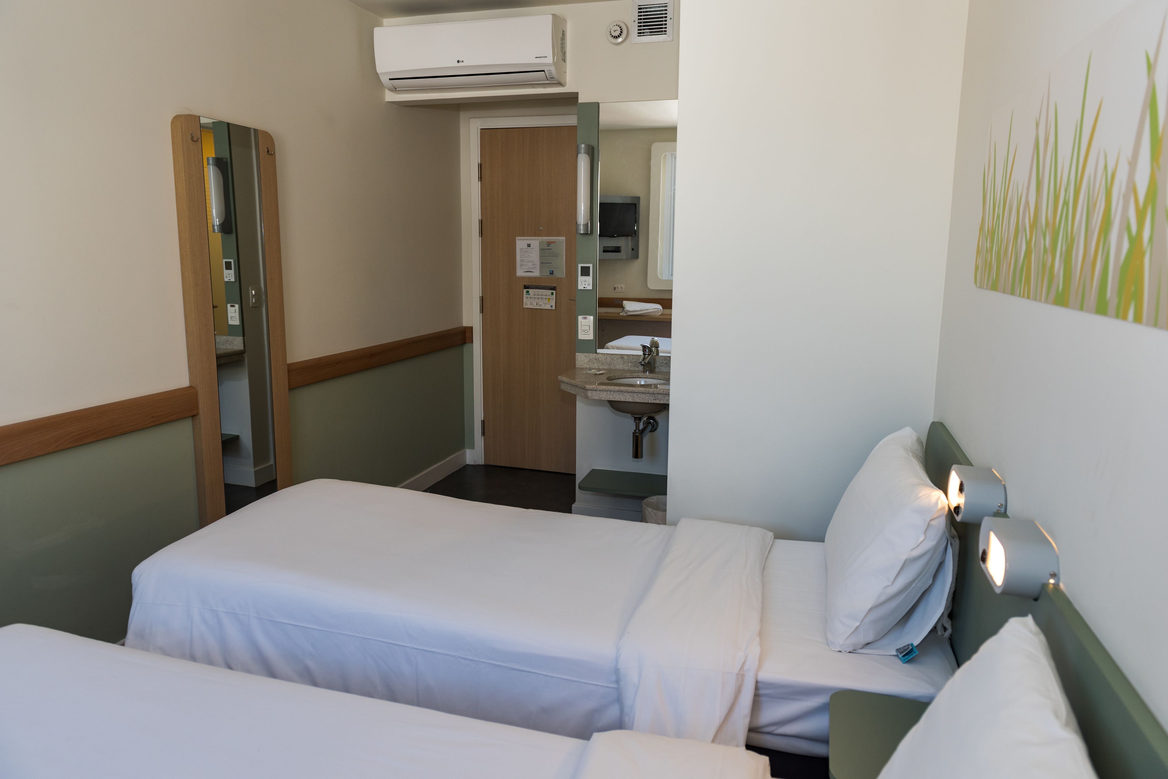 A hotel room with two single beds, a bathroom with a sink and mirror, and a hallway leading to another room.