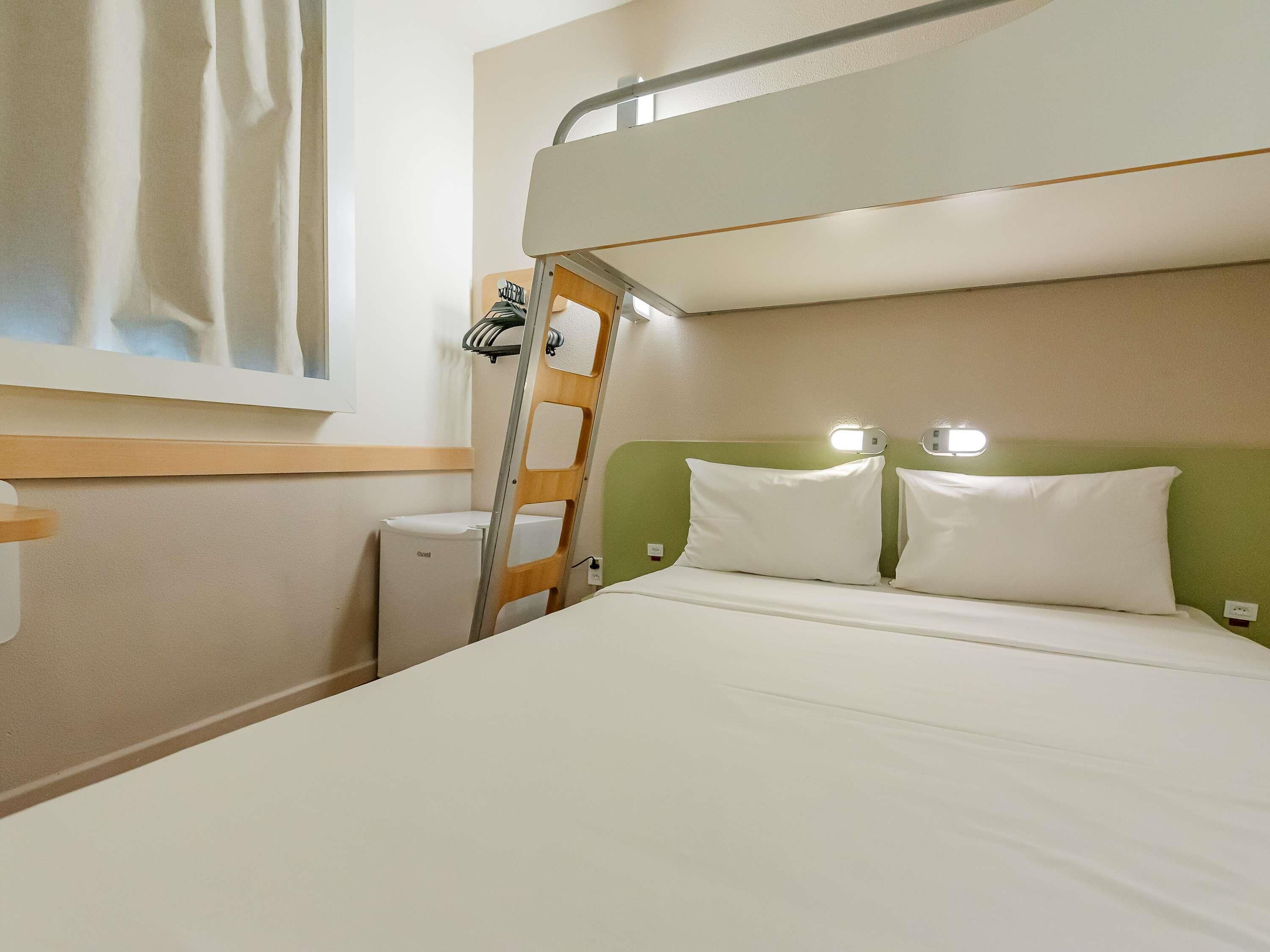 Una cama litera con escalera, una mesita de noche, una lámpara pequeña y una ventana con cortinas.