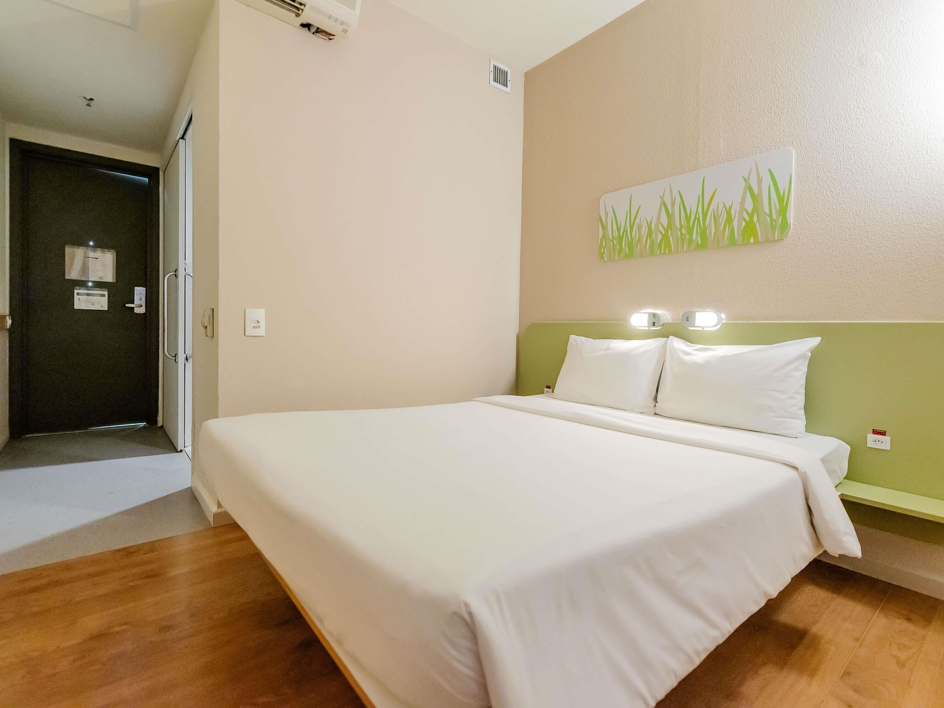 A hotel room with a bed, a door, a wall-mounted light, and a green accent wall.