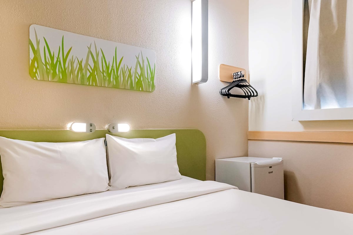 A hotel room with a bed, a green headboard, a nightstand, a small white cabinet, a wall-mounted towel rack, and a painting of grass.