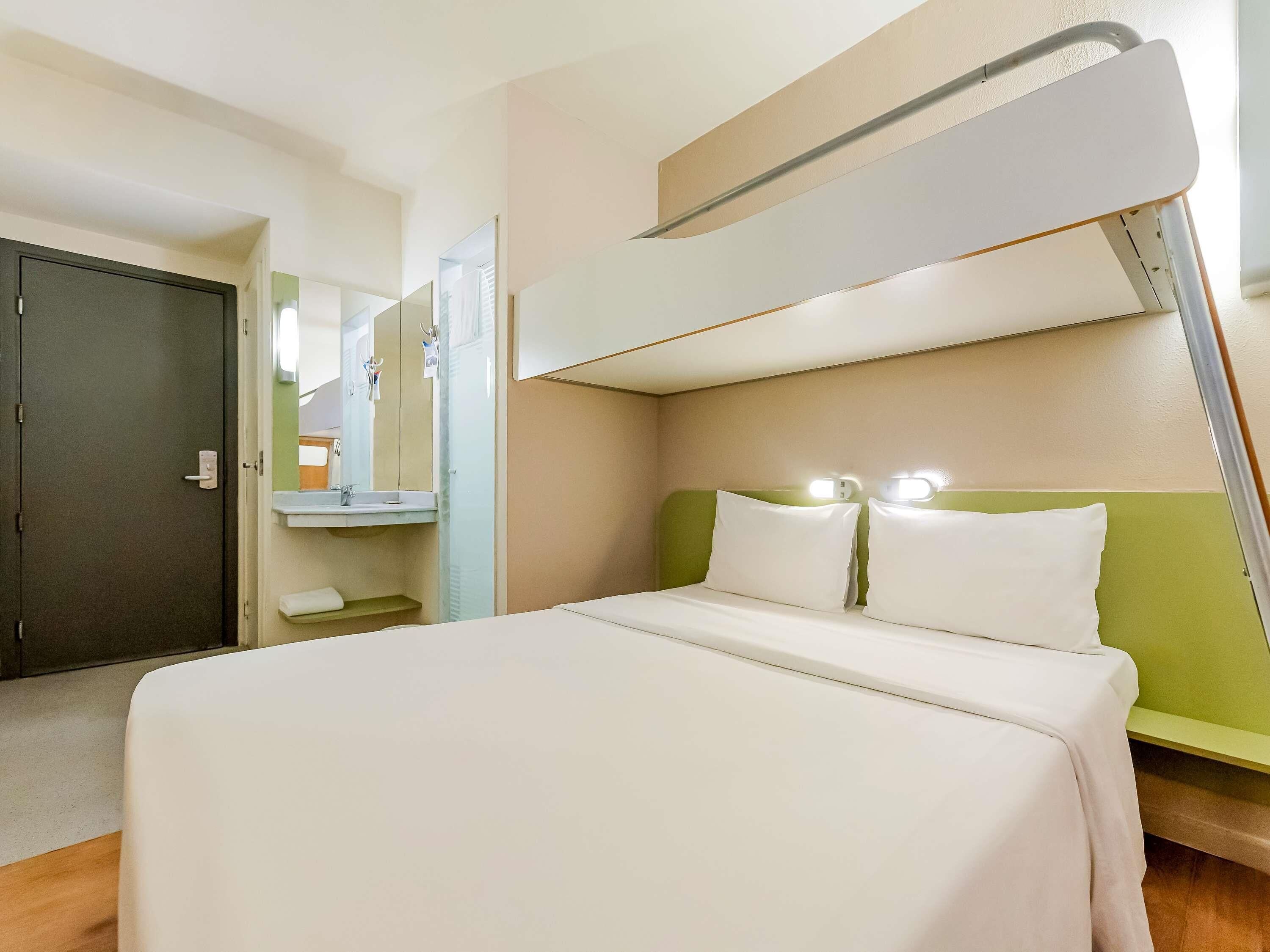 Habitación de hotel con literas, una mesita de noche, un espejo y un baño visible a través de una puerta abierta.