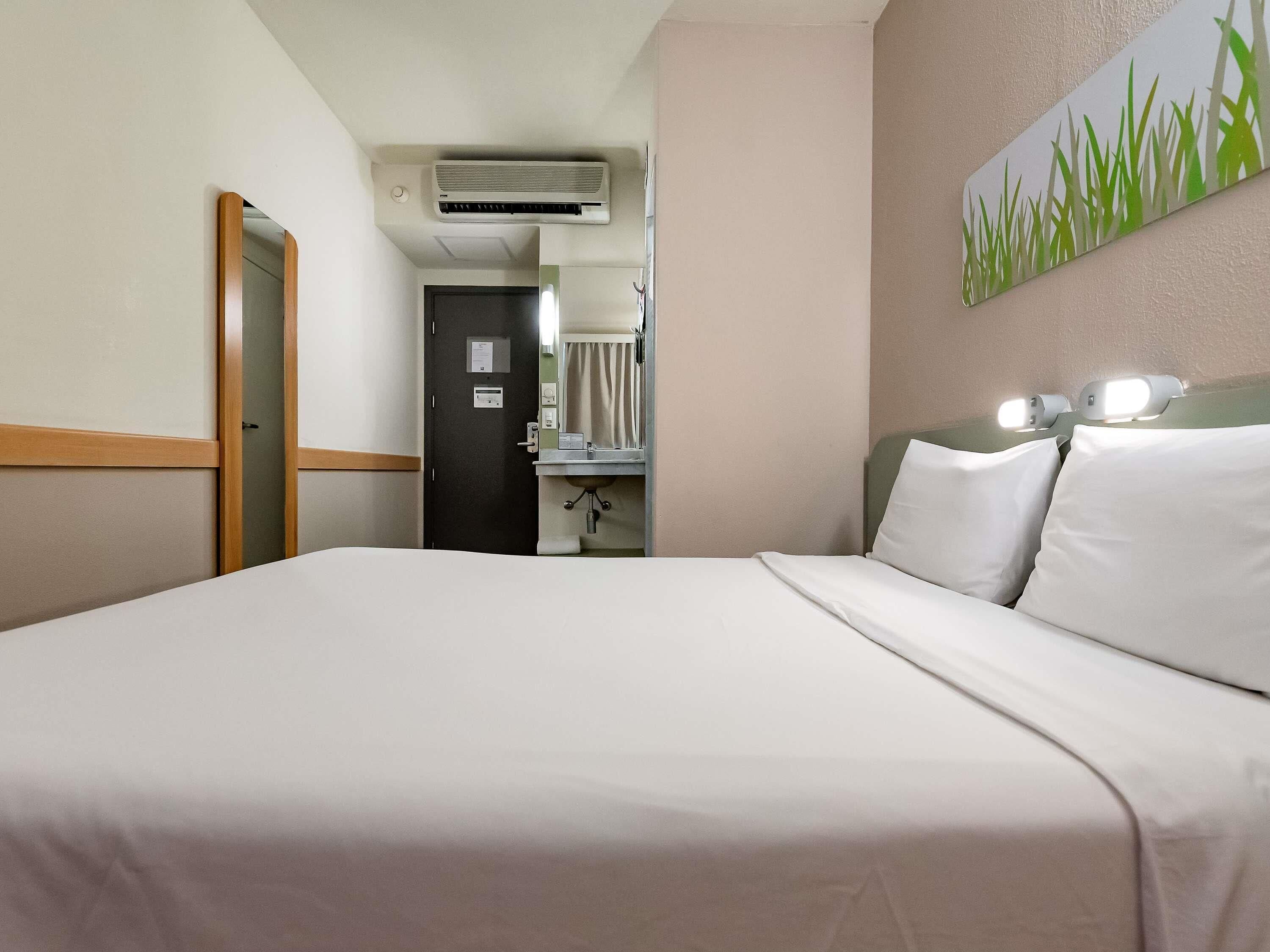 A hotel room with a bed, an air conditioning unit, a bathroom with a mirror, and a wall-mounted light fixture.