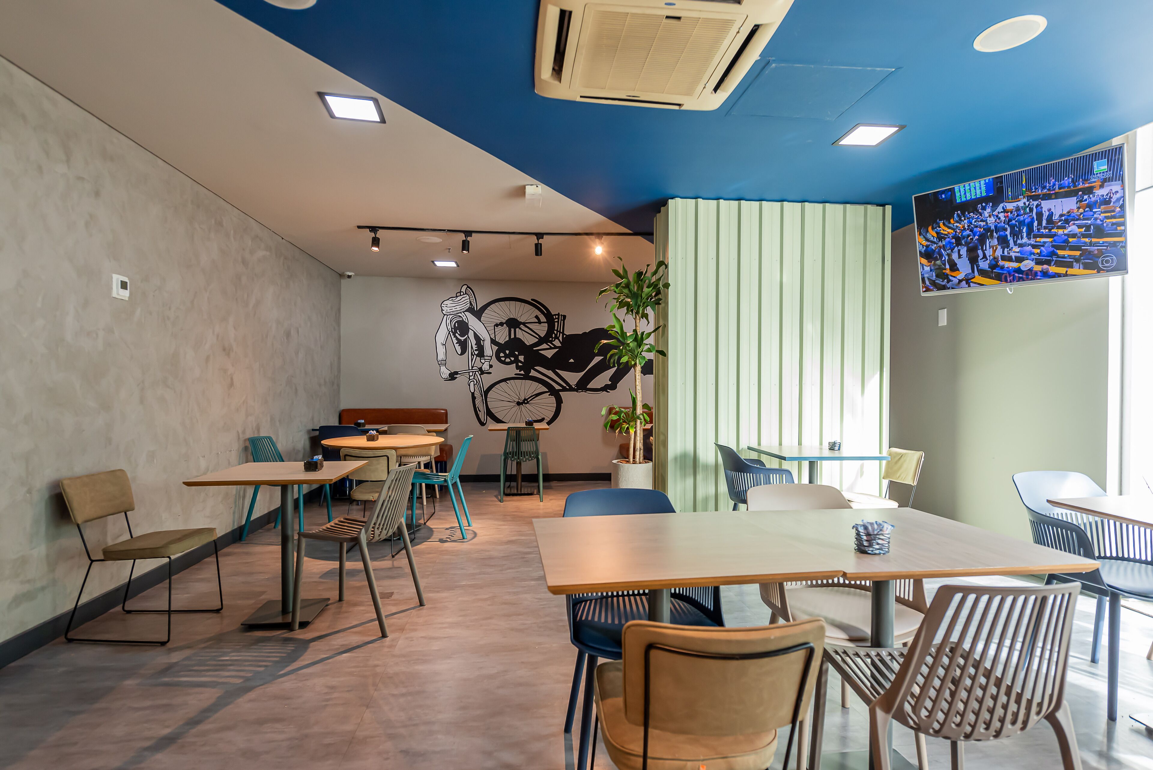 A modern cafe with tables and chairs, a wall-mounted TV, and a ceiling fan.
