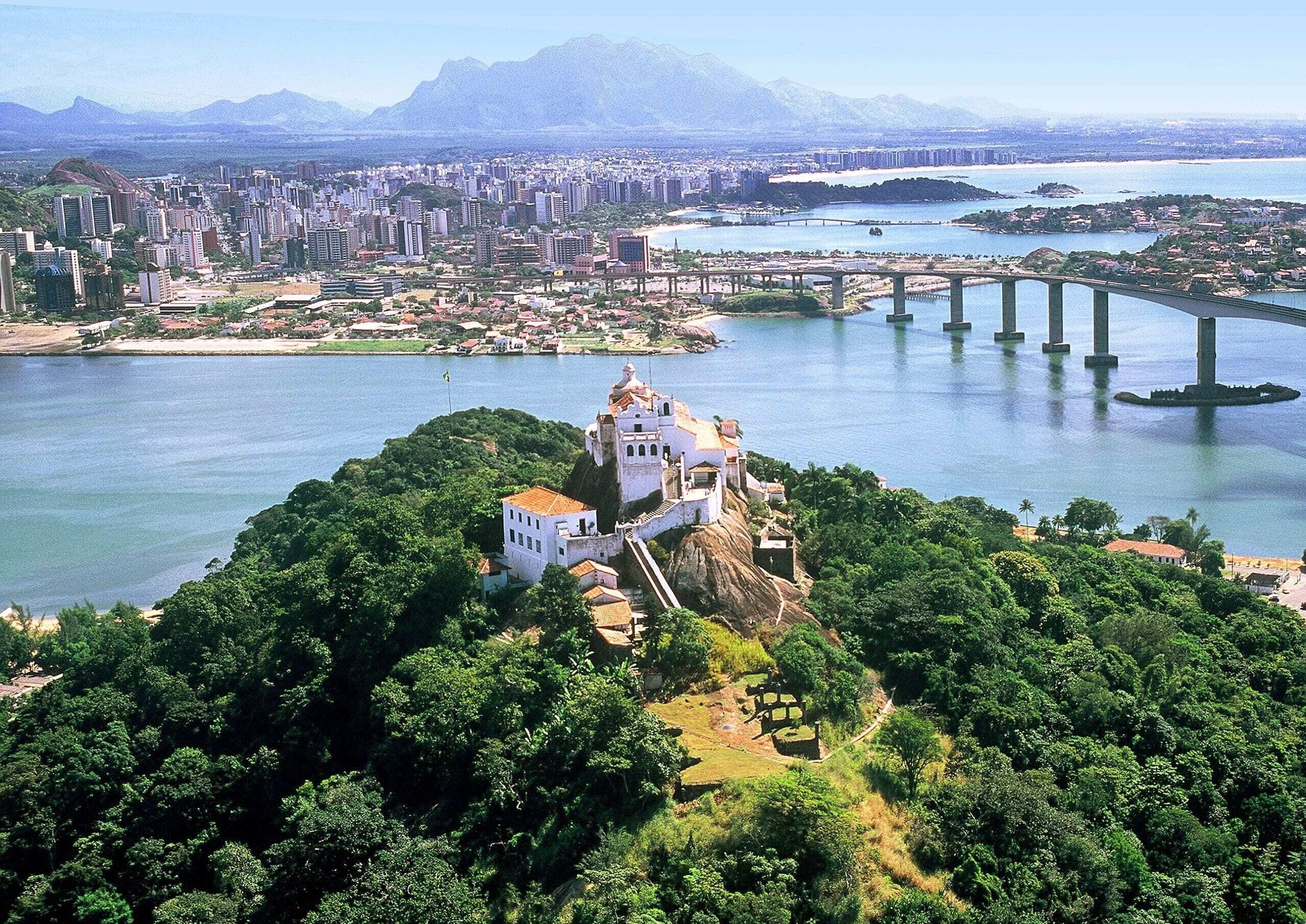 A historic building with a white facade, surrounded by lush greenery, overlooking a large body of water with bridges and a cityscape in the background.