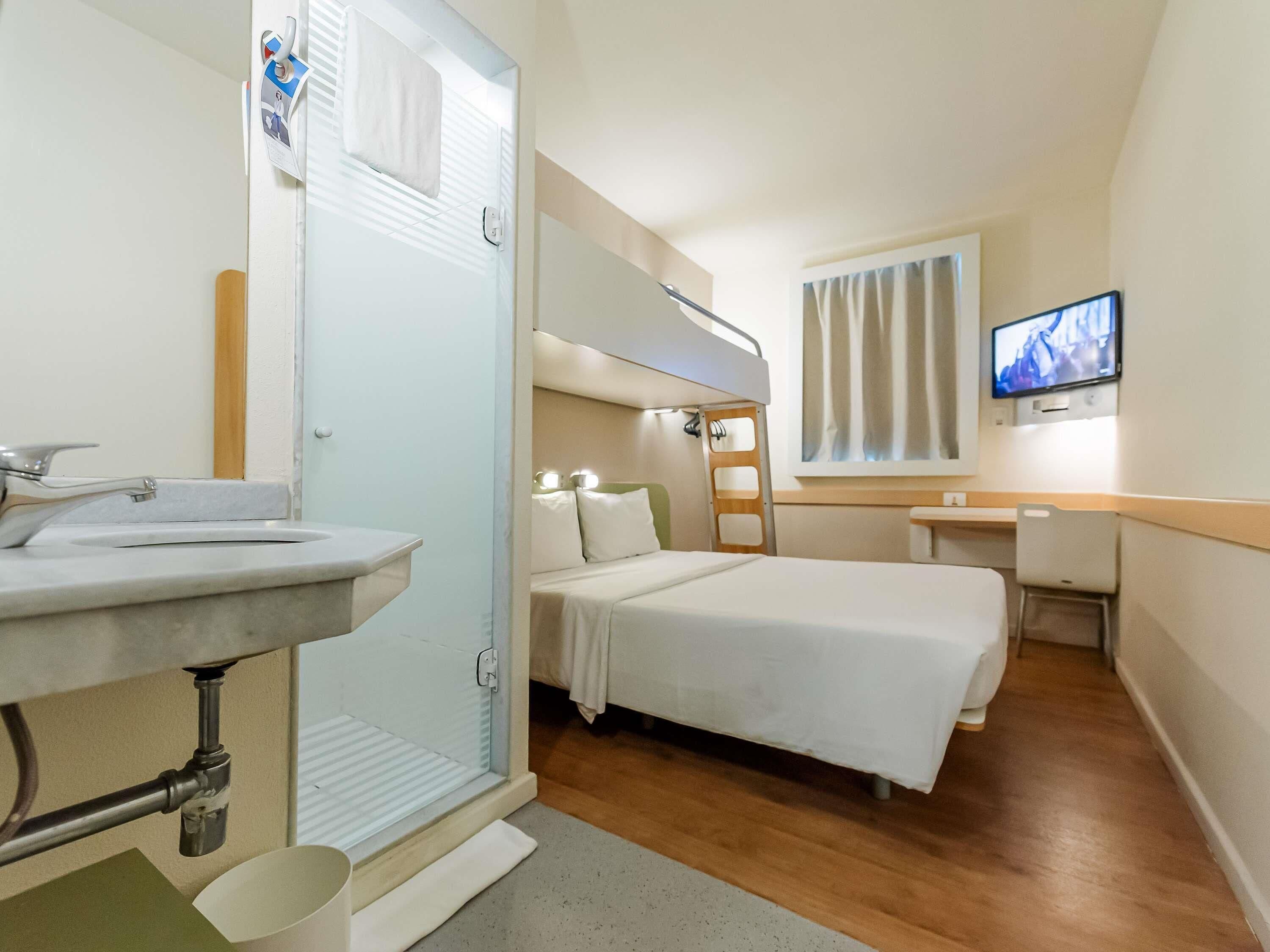 Habitación de hotel pequeña con literas, un escritorio, televisión y baño con lavabo y ducha.