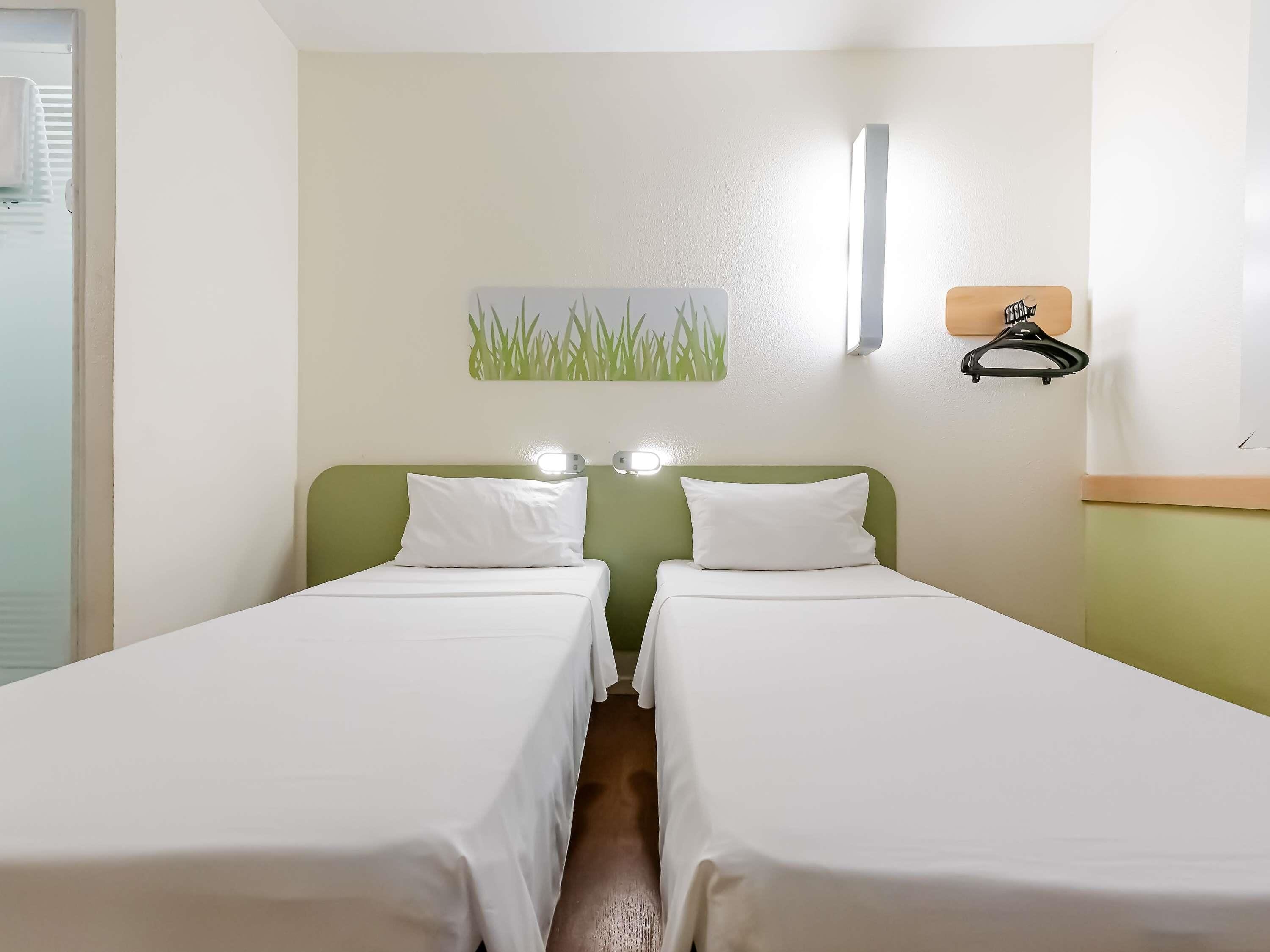 Ett hotellrum med två separata sängar, en grön sänggavel, en väggmonterad lampa och en liten tavla som föreställer gräs.