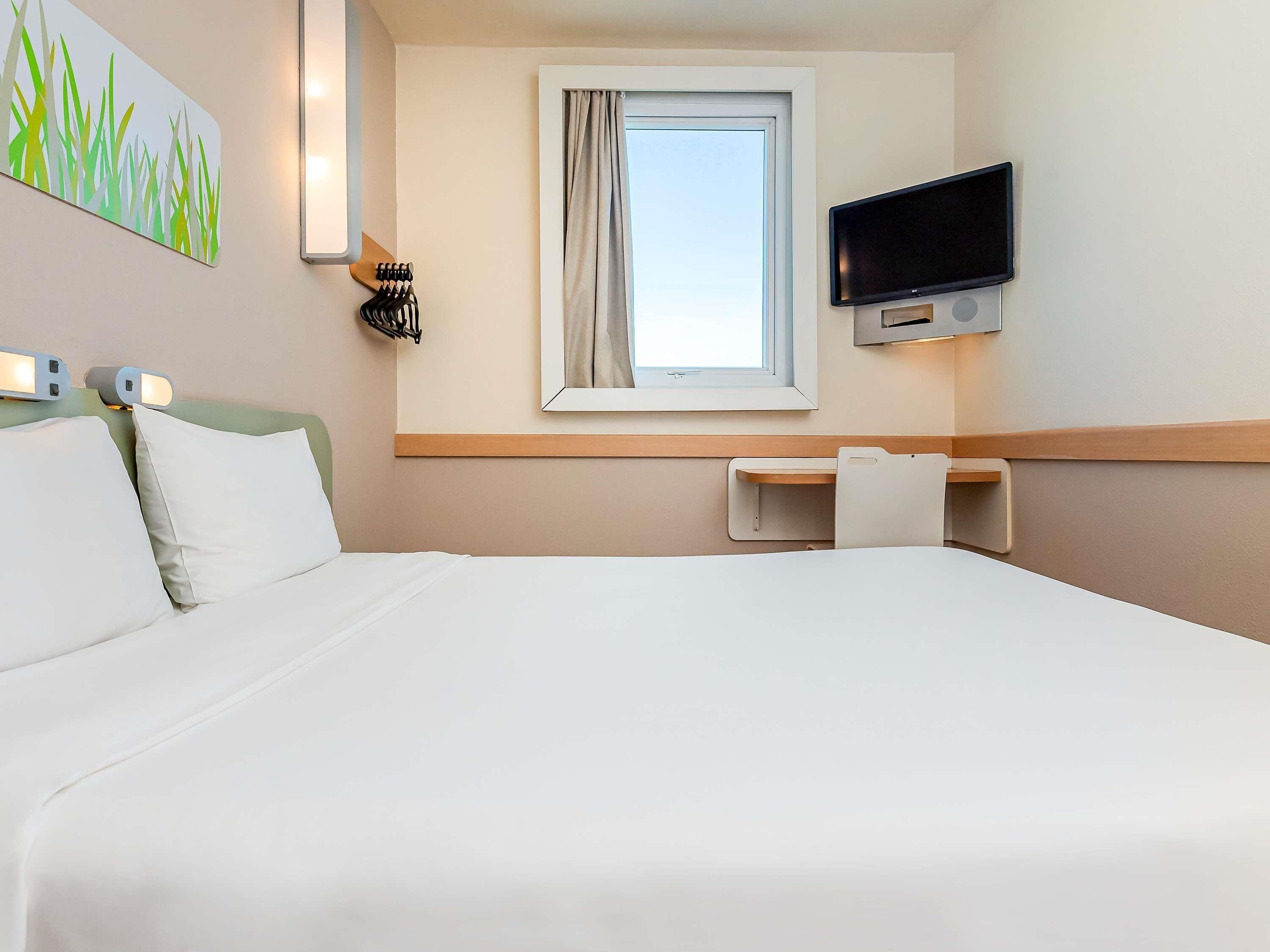 Una habitación de hotel con cama, escritorio, televisor y ventana con cortinas.