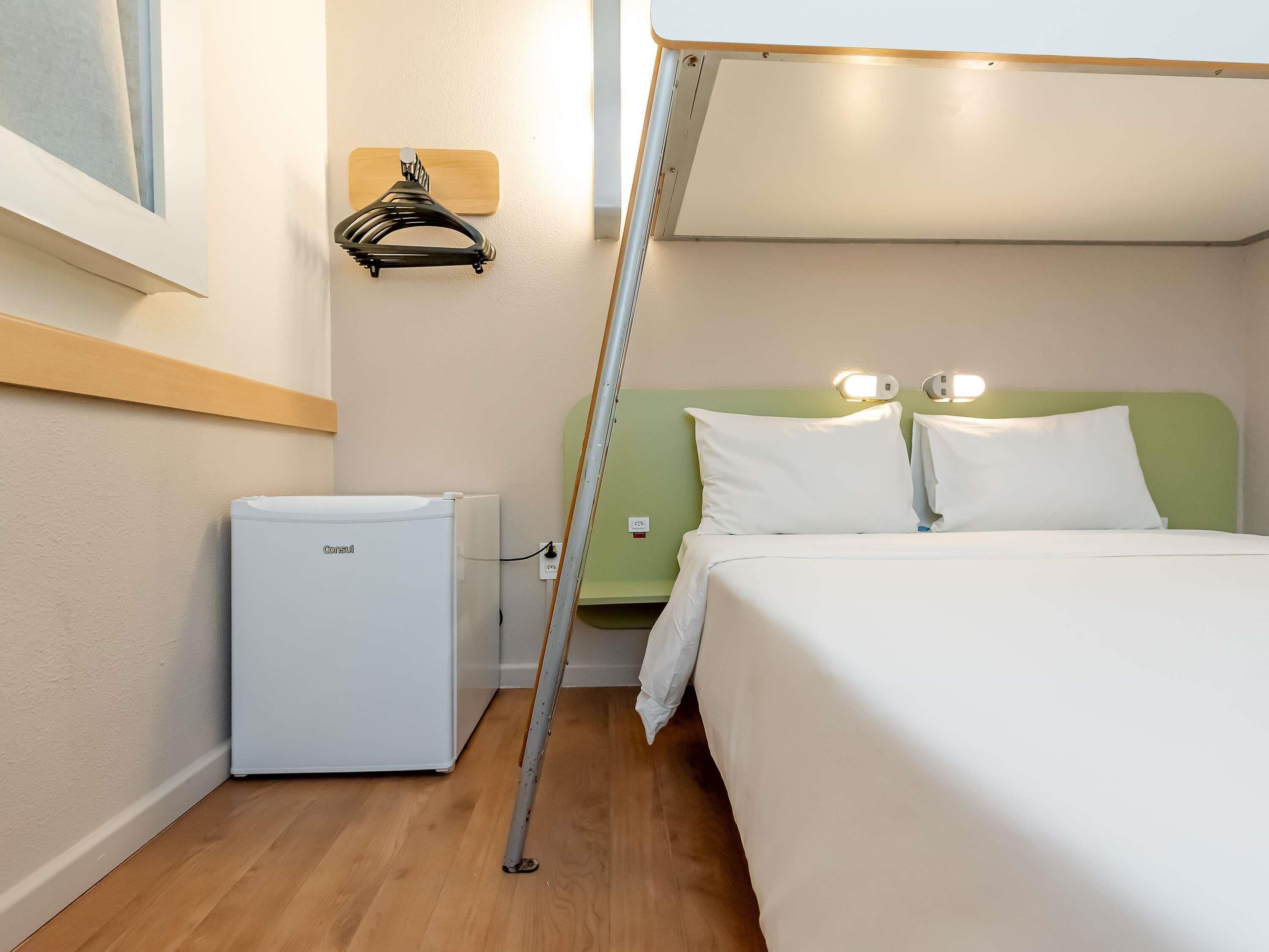 A compact hotel room with a bed, a small white refrigerator, a wall-mounted coat rack, and a green headboard.