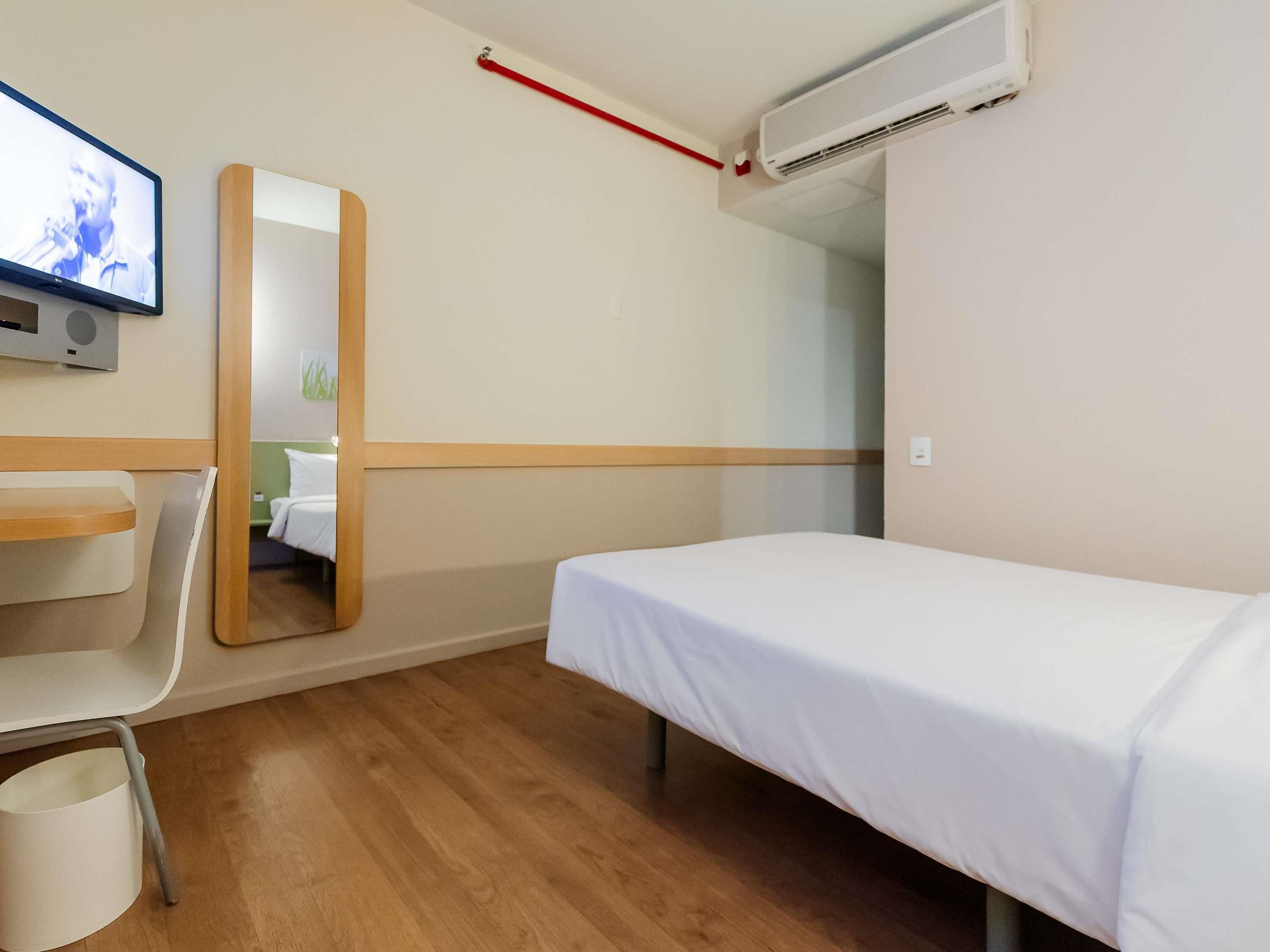 A hotel room with a single bed, a desk, a chair, a TV, and an air conditioning unit.