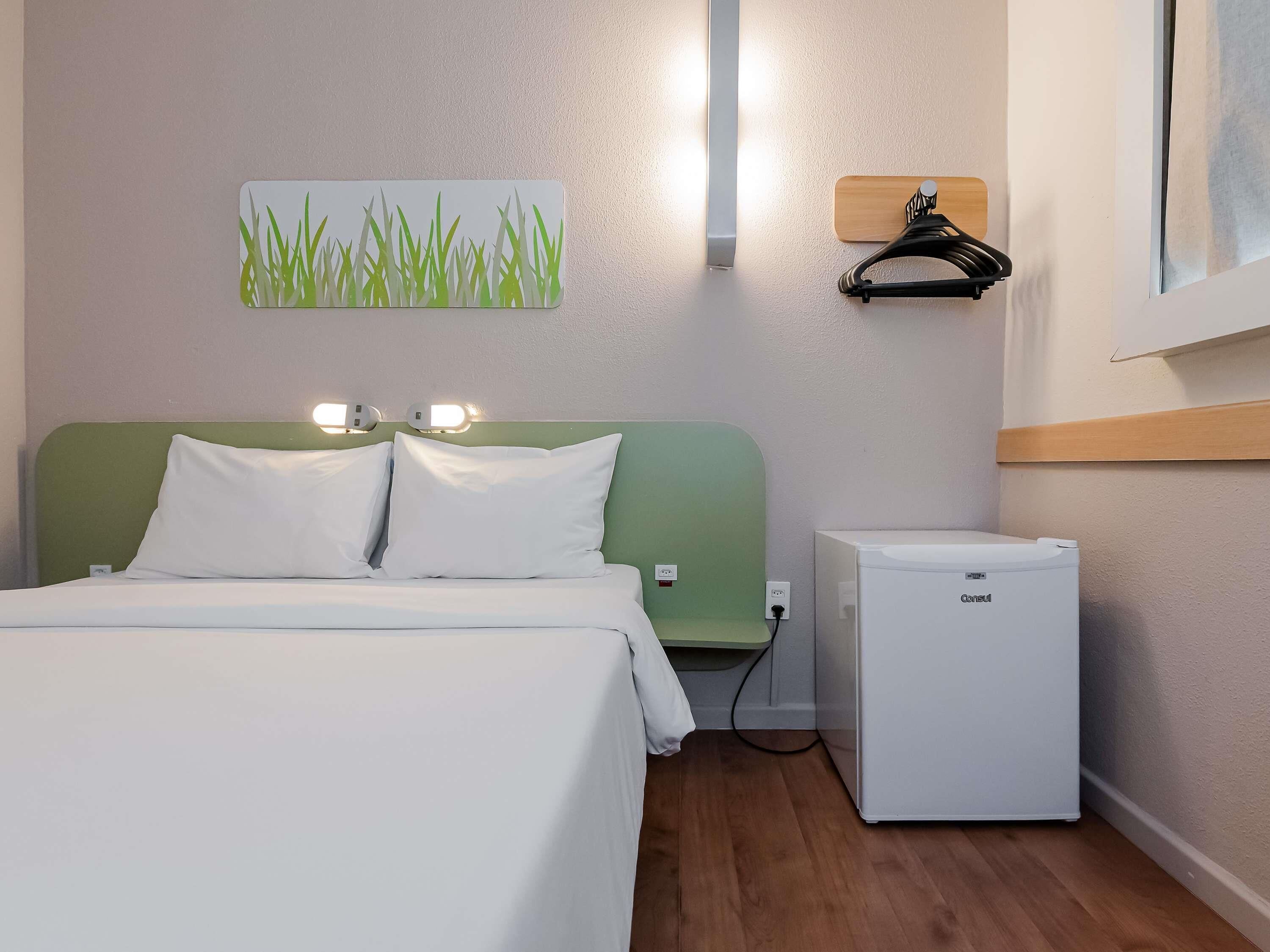 A hotel room with a bed, a small refrigerator, a wall-mounted heater, and a painting of grass.