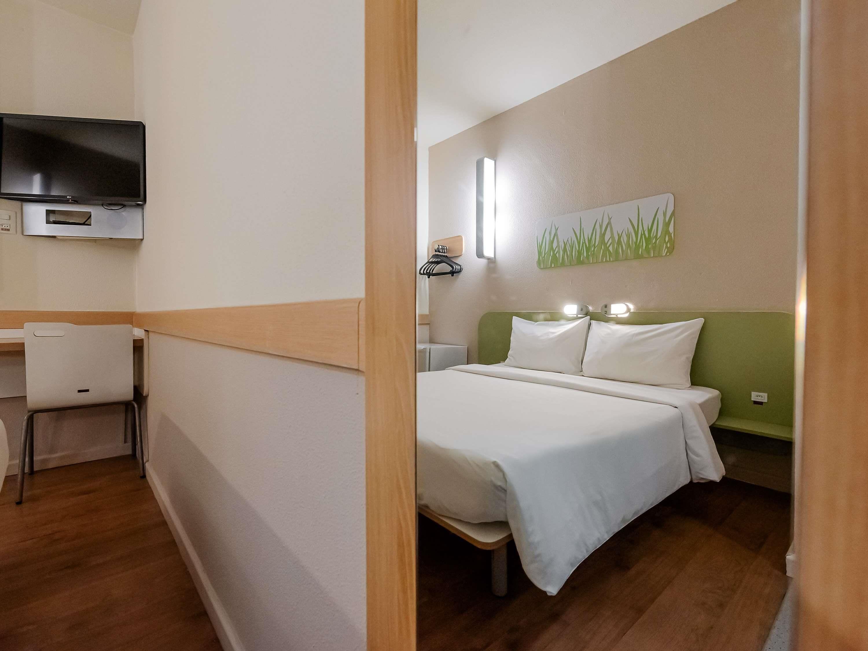 A hotel room with a bed, bedside table, a desk, a chair, a television, and a wall-mounted light fixture.