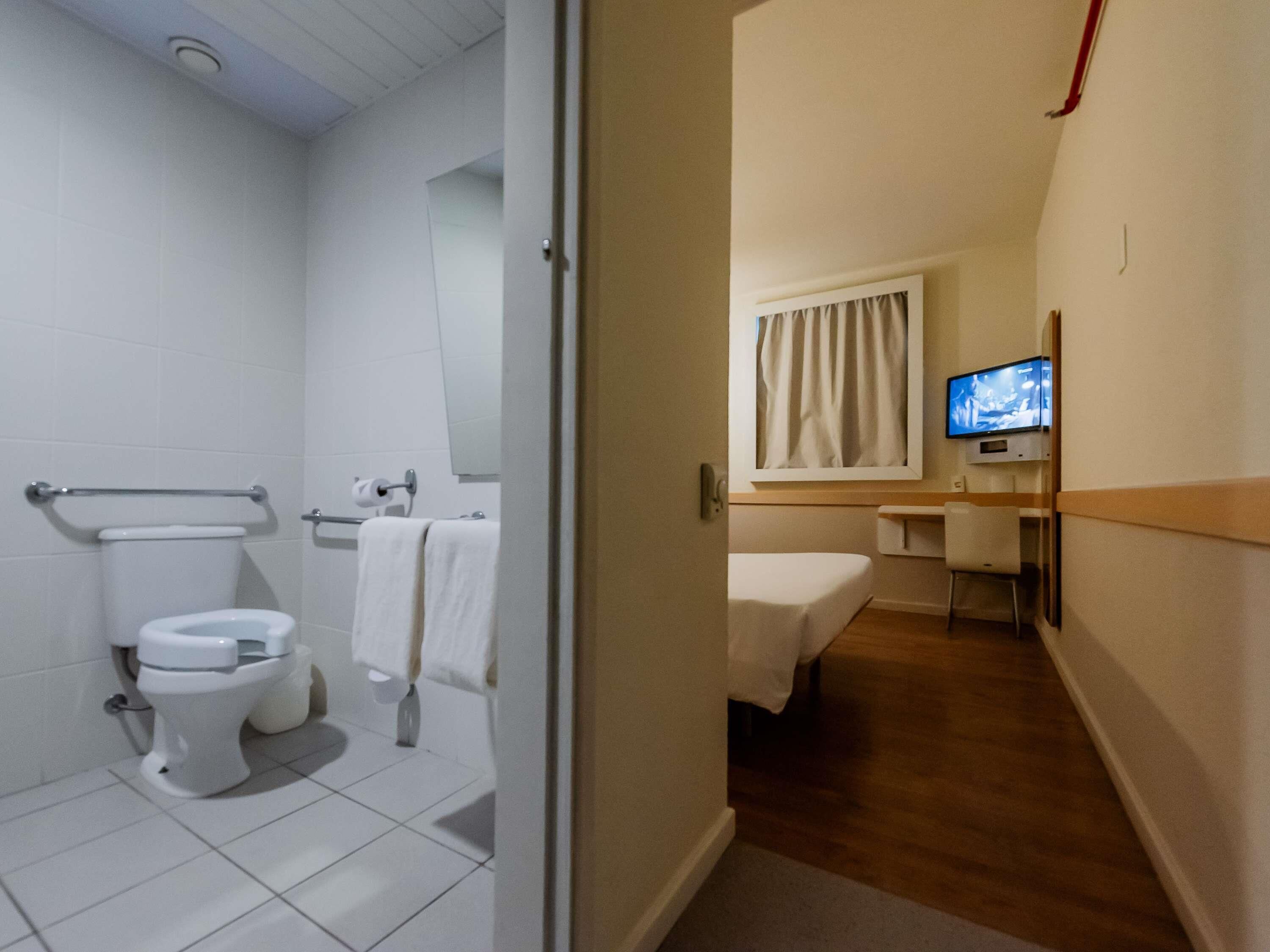 A hotel bathroom with a toilet, a shower, and a sink. There is a TV mounted on the wall and a window with curtains.