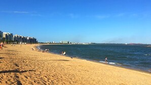 Une plage de sable fin, un ciel bleu dégagé, quelques personnes et des bâtiments en arrière-plan.