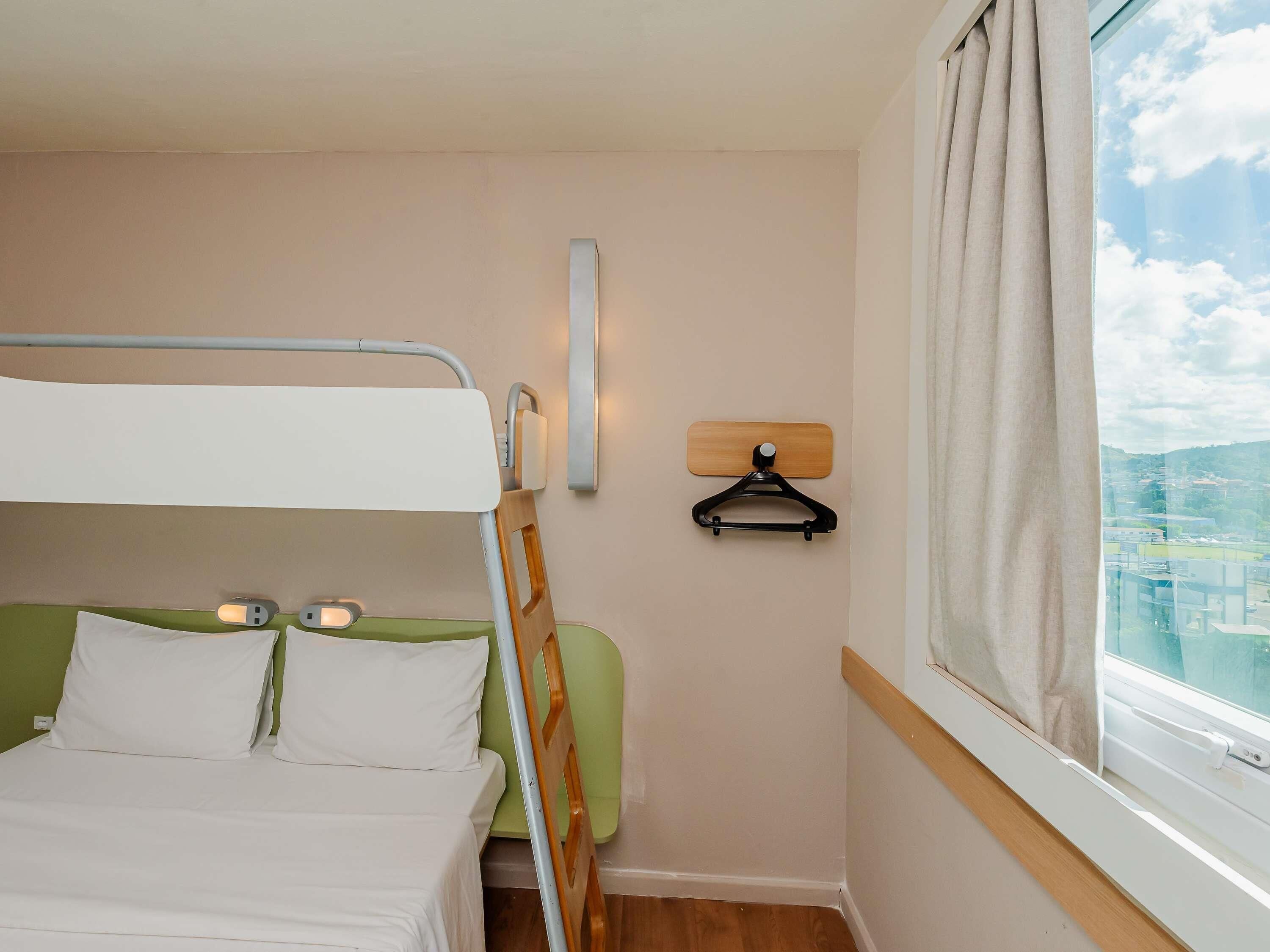 Un letto a castello con una testiera verde, un comodino con una lampada, una luce a parete e una finestra con vista sugli edifici.