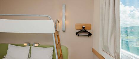 A bunk bed with a green headboard, a bedside table with a lamp, a wall-mounted light, and a window with a view of buildings.