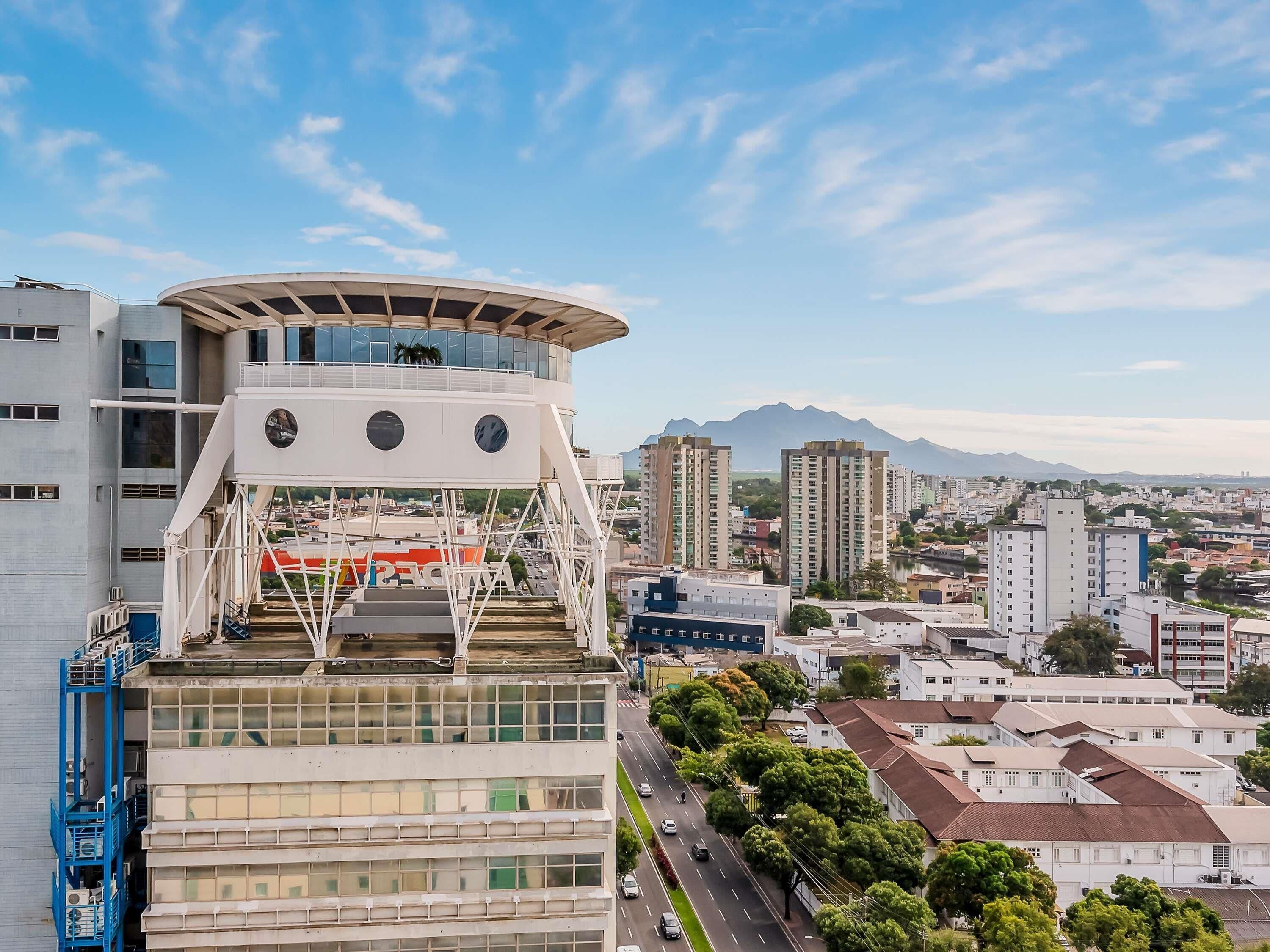Um edifício moderno com telhado curvo e grandes janelas, rodeado por um horizonte urbano com edifícios residenciais altos e uma montanha ao fundo.