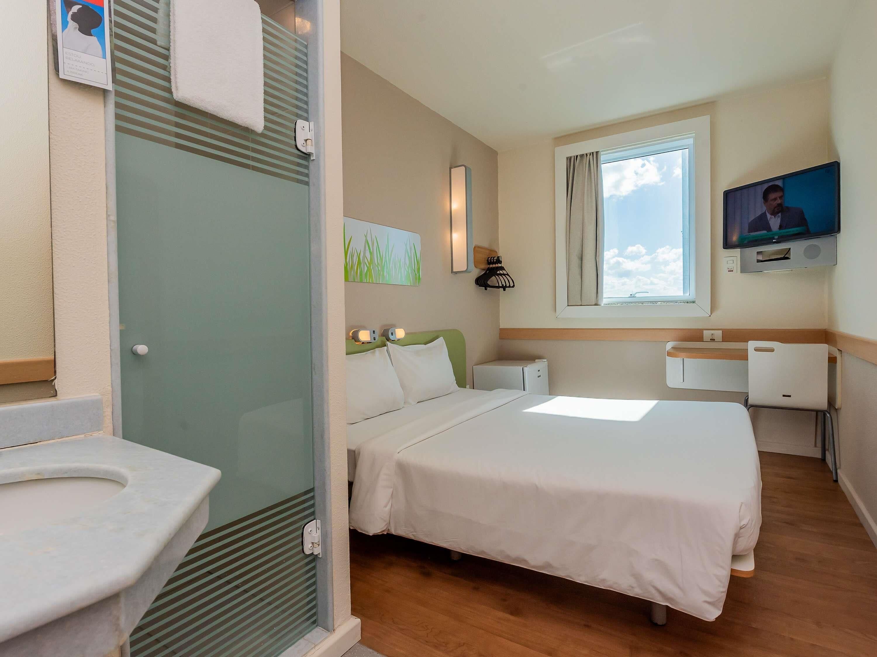 A hotel room with a bed, bedside table, lamp, TV, window, and a bathroom with a sink and toilet.