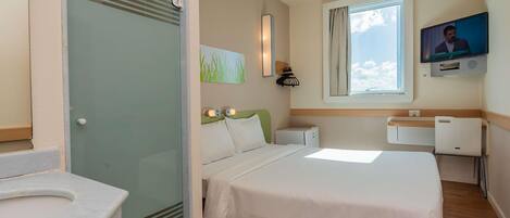 A hotel room with a bed, bedside table, lamp, TV, window, and a bathroom with a sink and toilet.