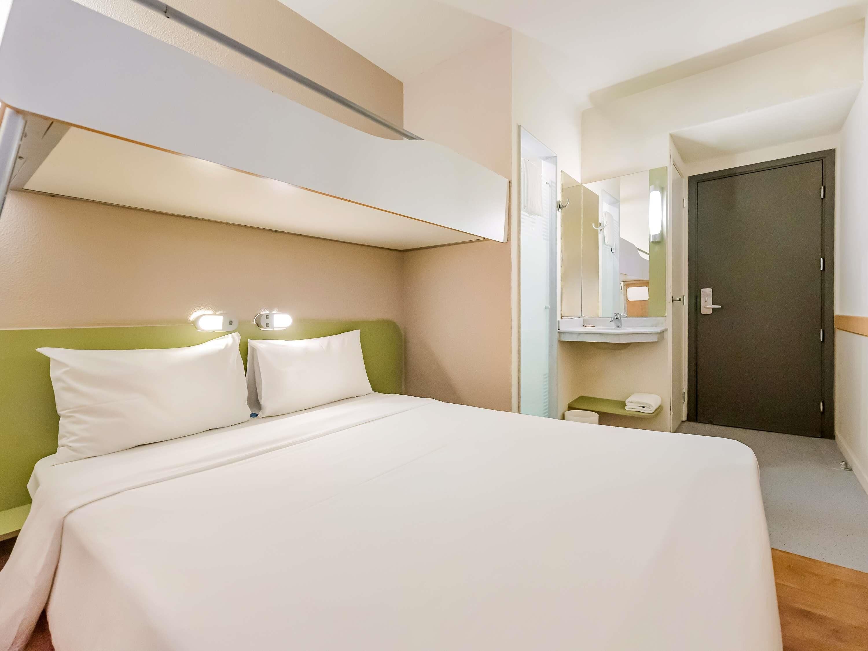 A hotel room with a bed, a bedside table, a mirror, a door, and a small shelf with towels.