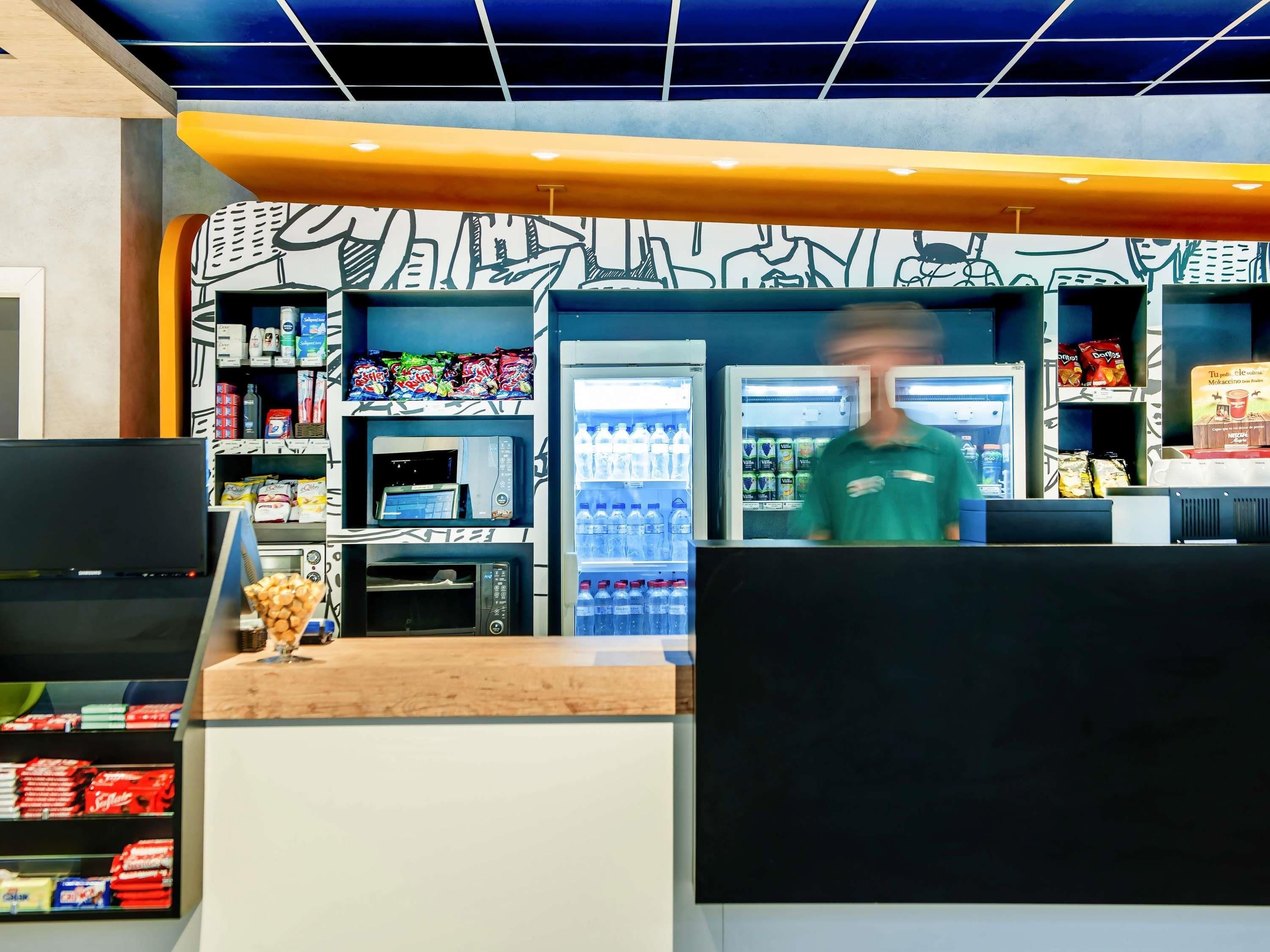 A fast-food restaurant interior with a counter, a cash register, a refrigerator stocked with beverages, and shelves with snacks.