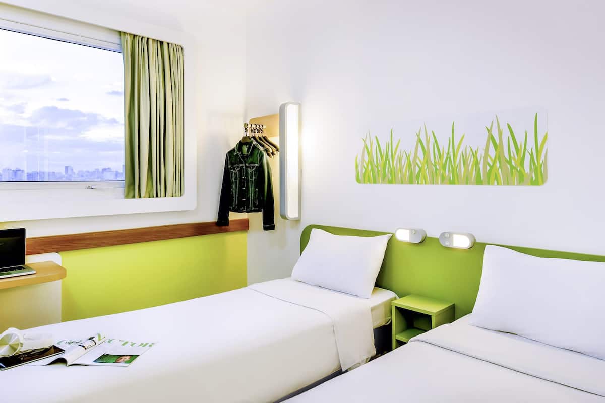 A hotel room with two beds, a desk with a laptop, a window with curtains, and a wall art of grass.