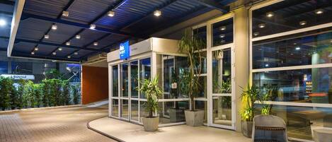 A modern building entrance with glass doors, potted plants, and a sign indicating a restaurant.