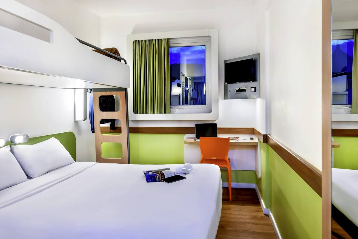 A hotel room with a bed, a desk with a chair, a TV, and a window with curtains.