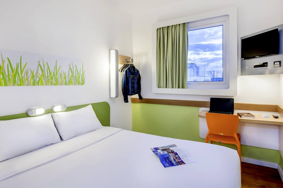 A hotel room with a bed, a desk with a laptop, a TV, a window with curtains, and a wall with a grass art piece.