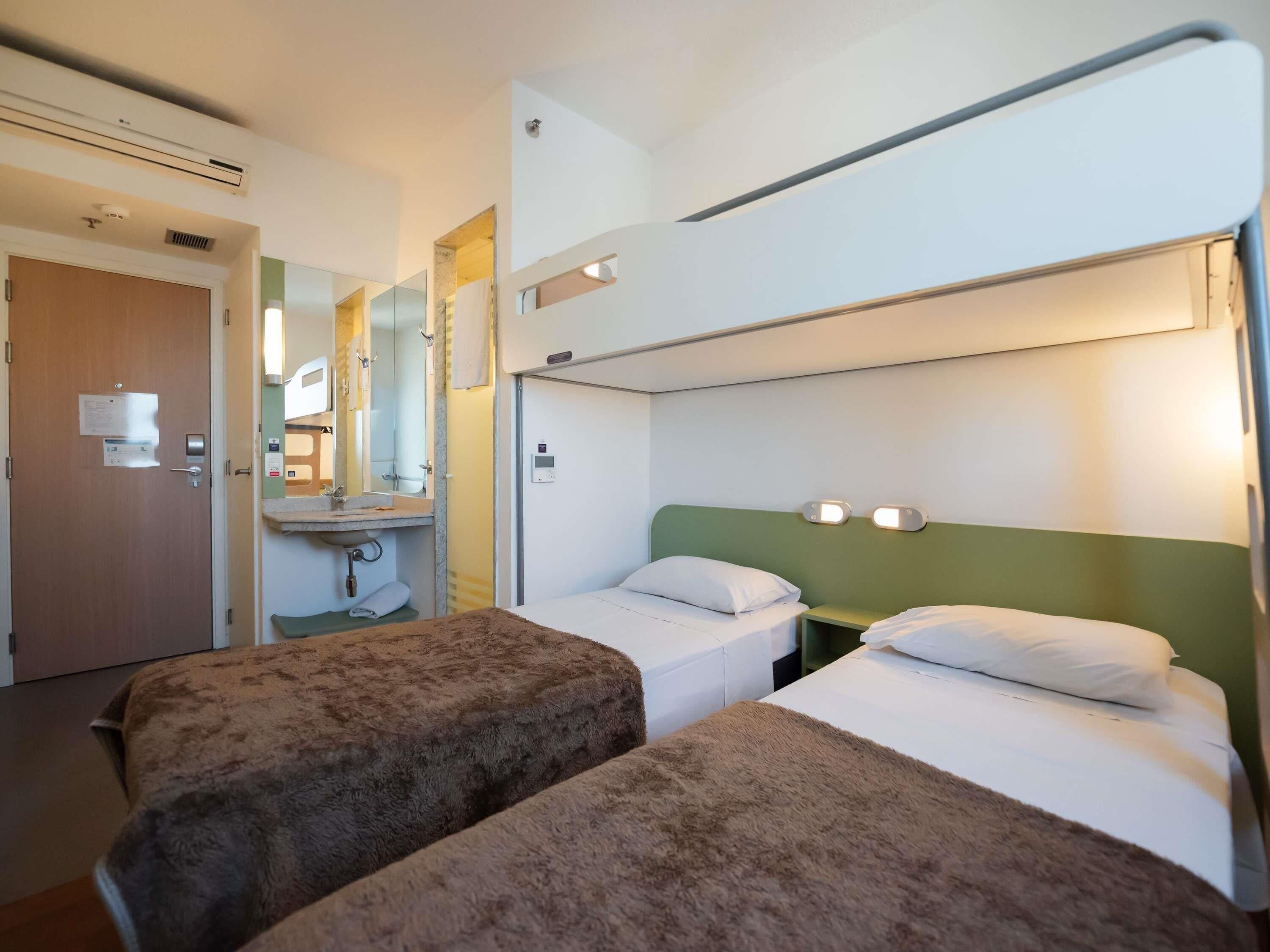 Quarto de hotel com duas camas, banheiro com pia e espelho, e aparelho de ar condicionado.