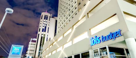Un hotel moderno con un llamativo letrero de 'ibis budget', fachada de vidrio y un área de entrada pavimentada.