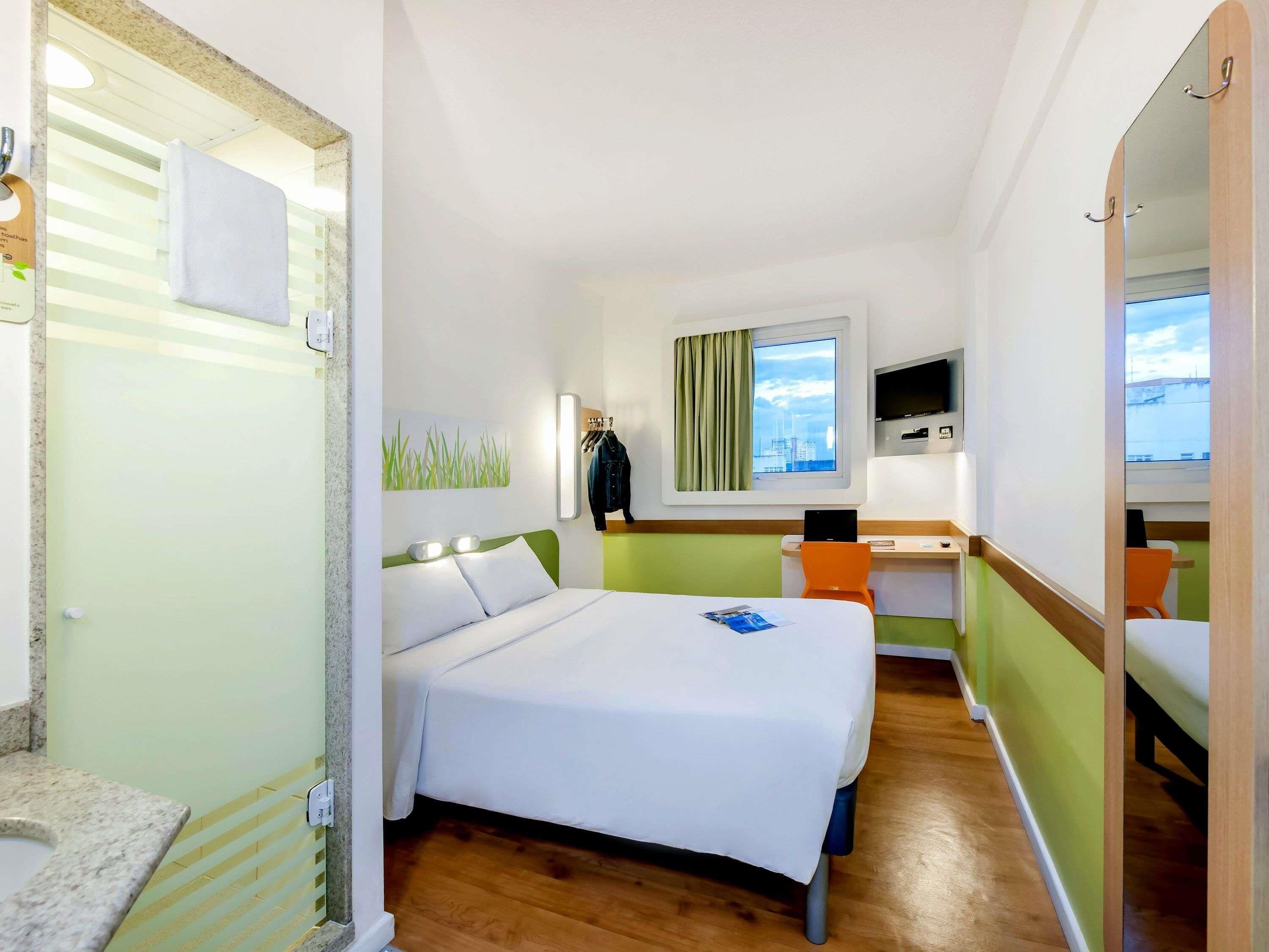 Quarto de hotel com cama, escrivaninha, cadeira, televisão e janela com vista para a cidade.