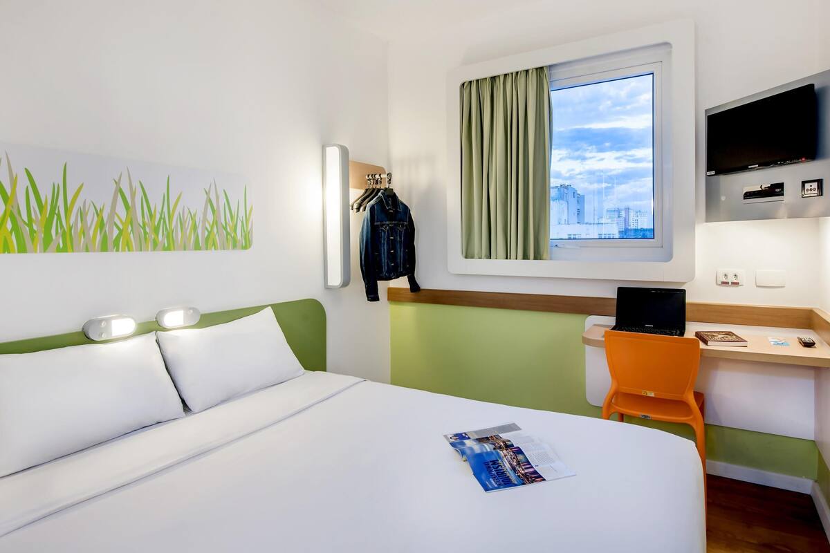A hotel room with a bed, a desk with a chair, a TV, a window with curtains, and a wall with a plant decoration.