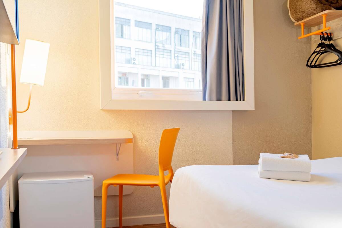 Une chambre d’hôtel avec un lit, une chaise, une petite table, une lampe et une fenêtre donnant sur des immeubles.
