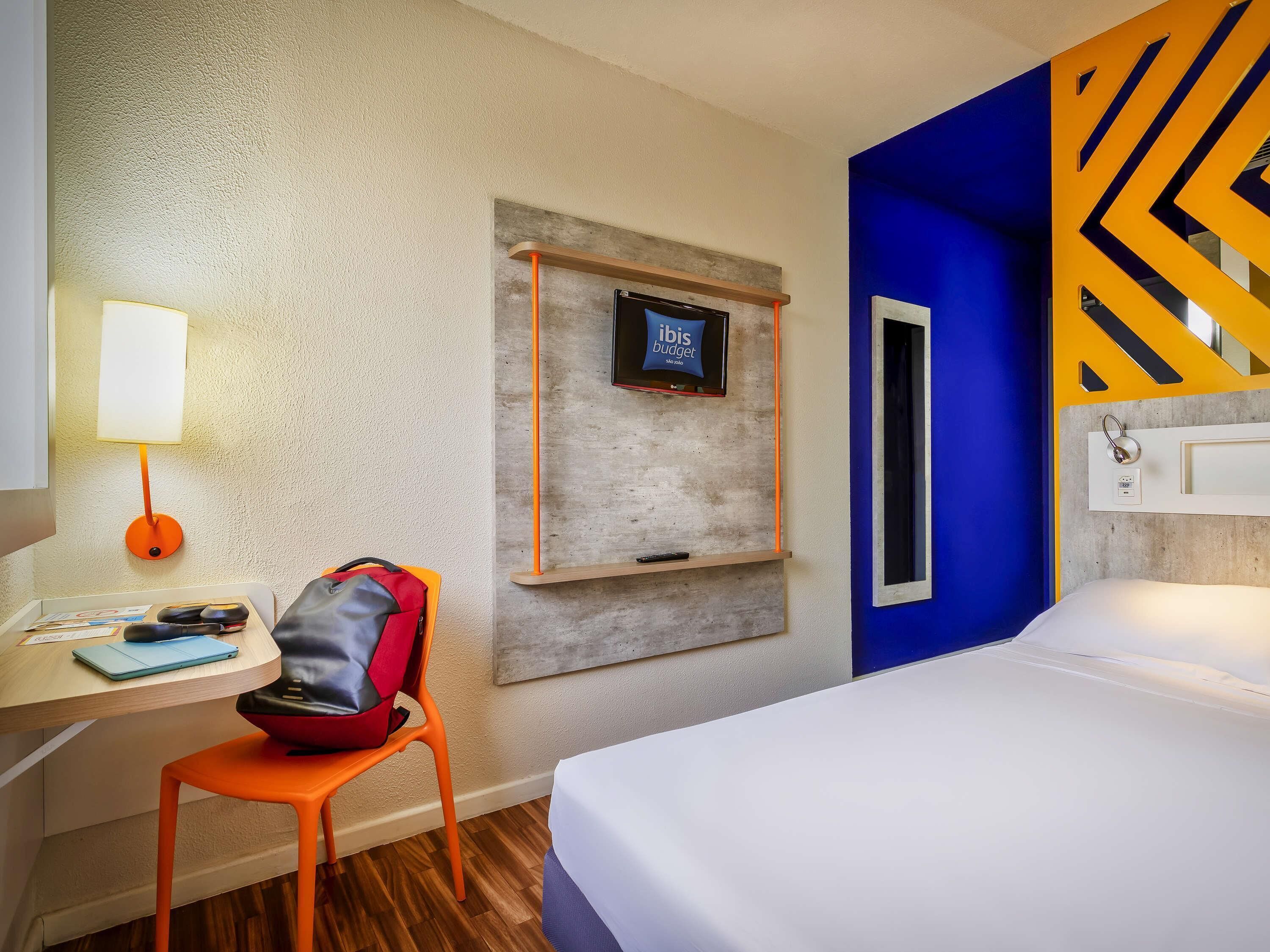 Habitación de hotel con una cama, un escritorio con lámpara, una silla, un televisor montado en la pared y una pared de acento azul.