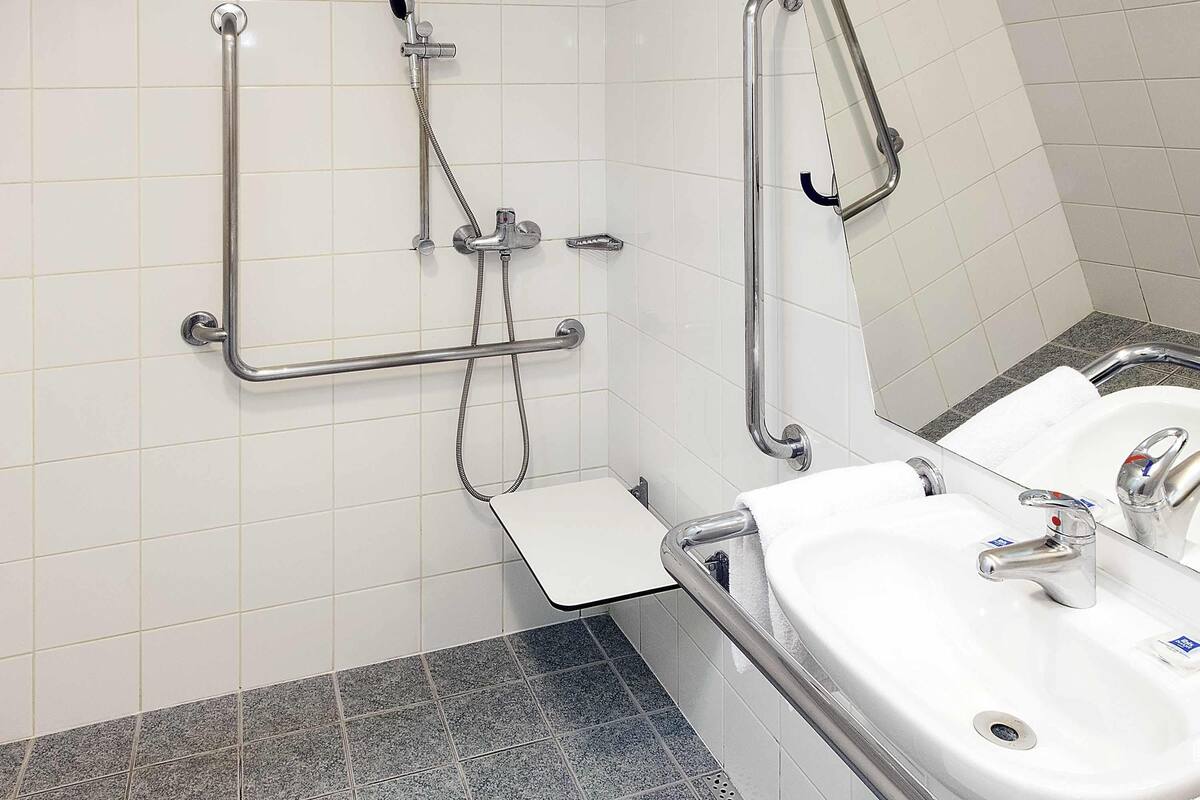 Une salle de bain avec deux lavabos, un miroir, une douche équipée de barres d’appui et un sol carrelé.