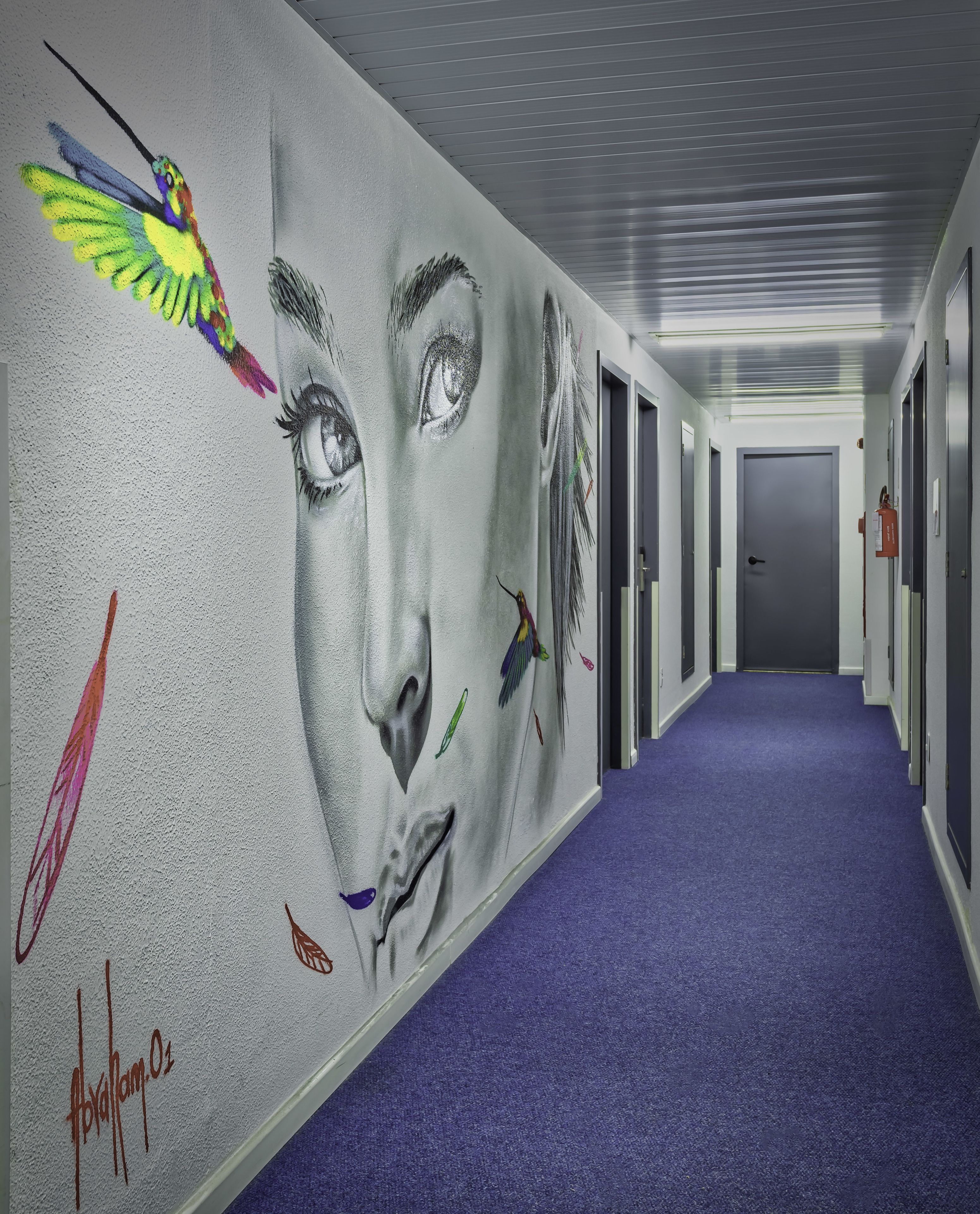 A hallway with a large mural featuring a bird and a human face. The hallway has a purple carpet and doors on the right.