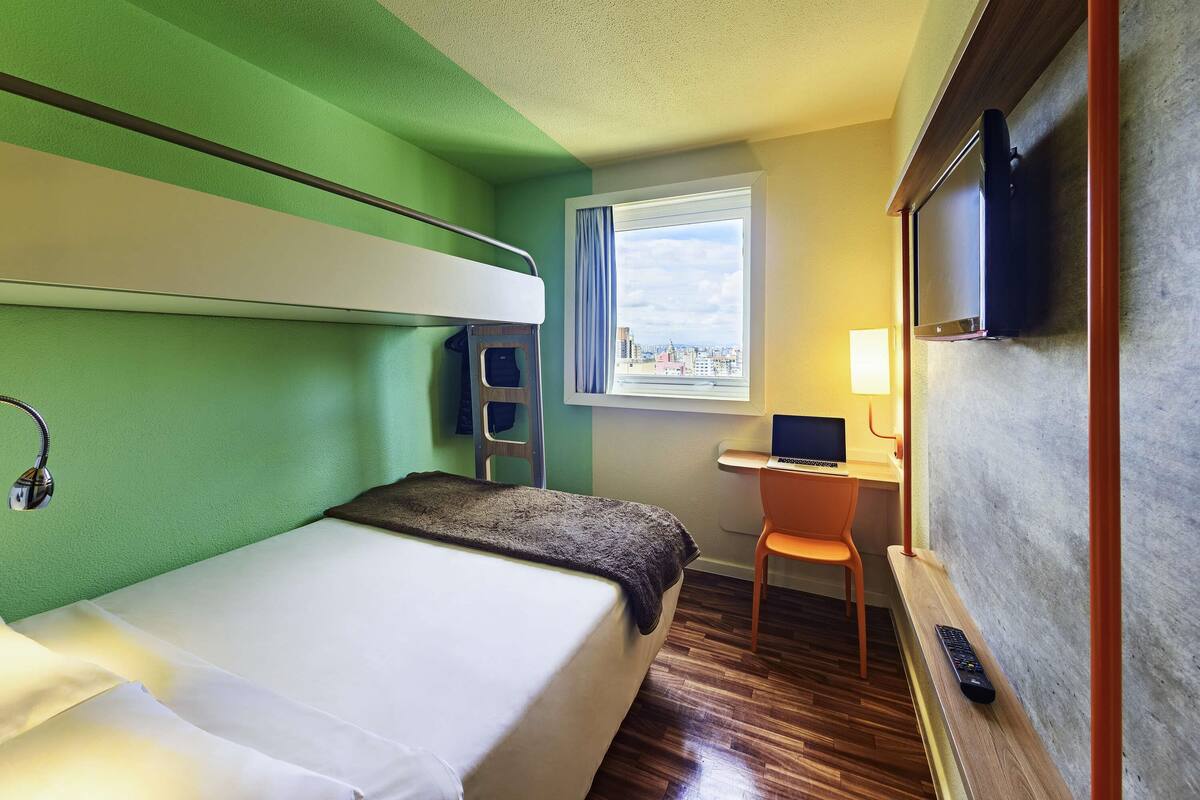 Une chambre d’hôtel avec un lit superposé, un bureau avec un ordinateur portable, une chaise, une télévision et une fenêtre donnant sur la ville.