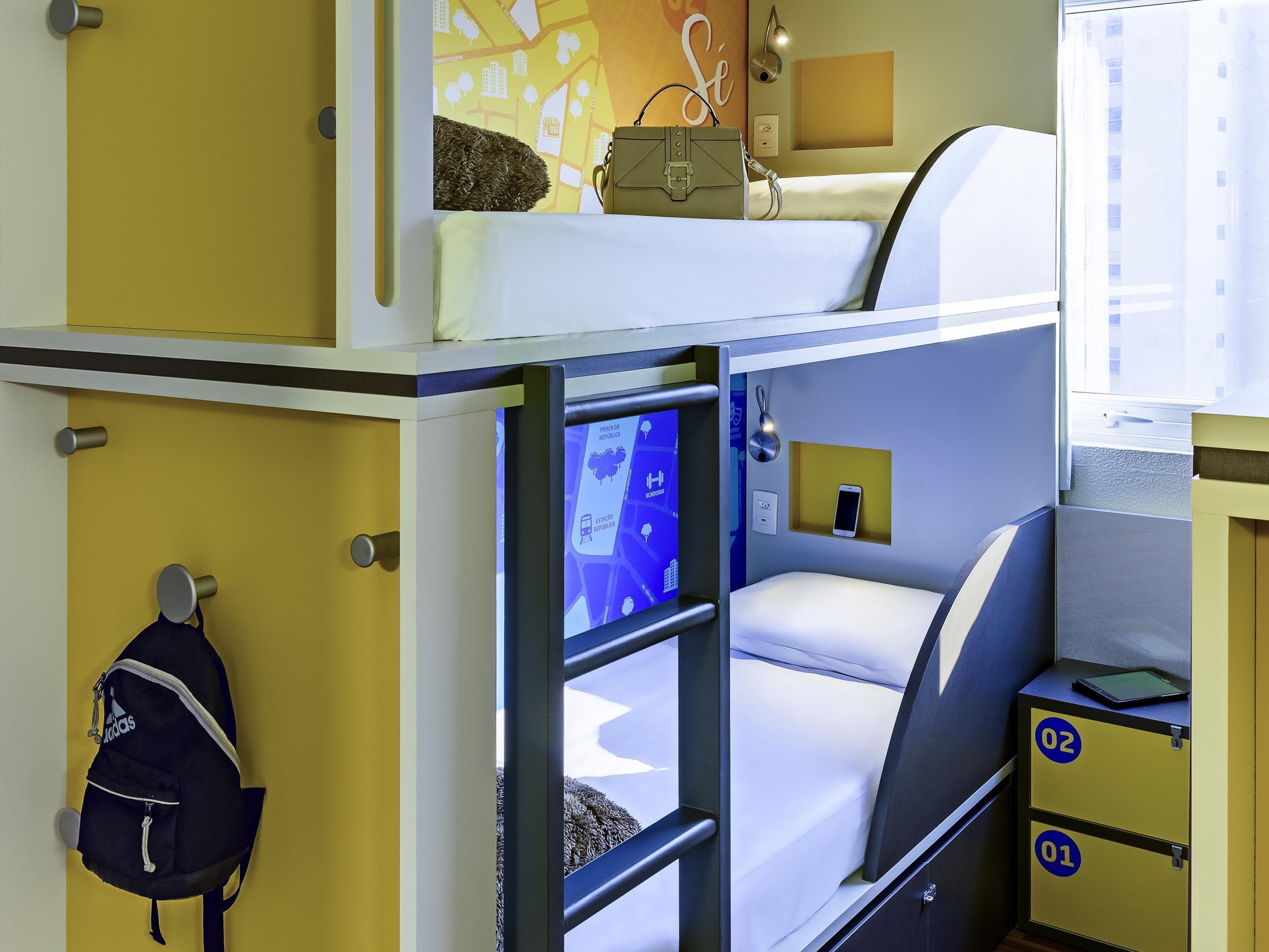 Una habitación de hotel compacta con literas, un escritorio pequeño con una tableta y una estación de carga en la pared.