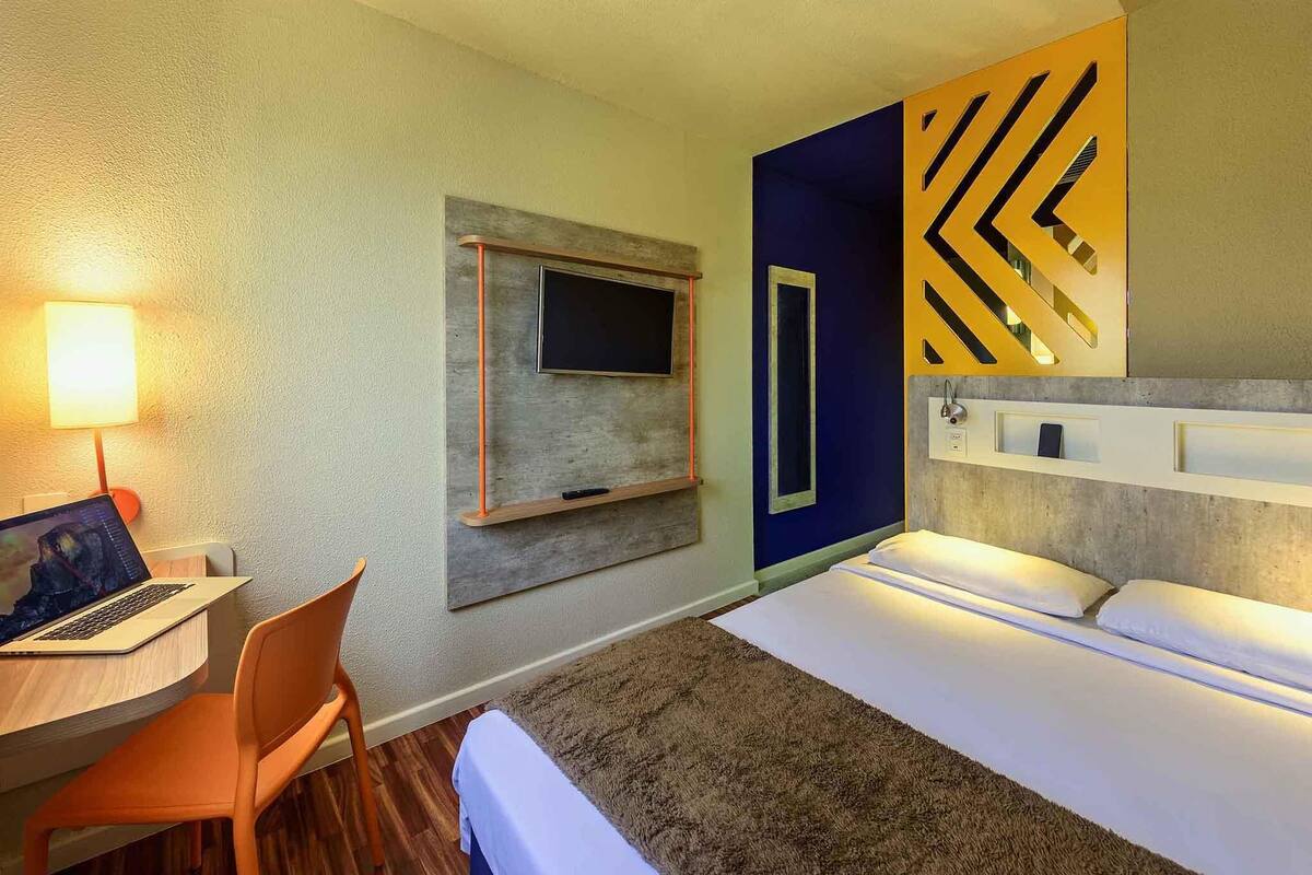 Une chambre d’hôtel comprenant un lit, un bureau avec un ordinateur portable, une chaise, un téléviseur fixé au mur et un panneau décoratif mural.