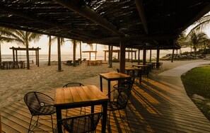 A beachside patio with wooden tables and chairs, a thatched roof, and a view of the ocean.