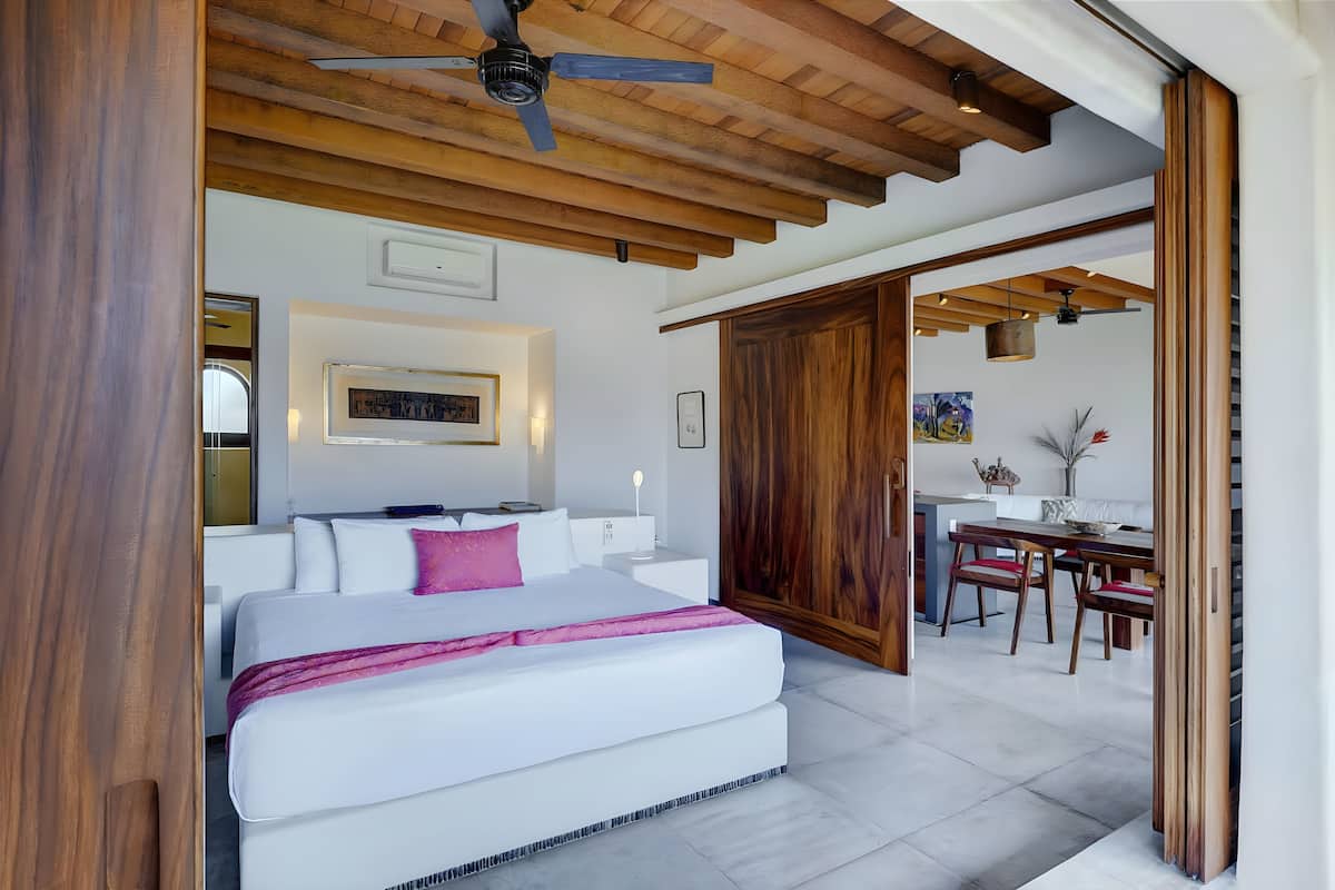 Une chambre moderne avec un grand lit, des poutres apparentes en bois au plafond et un coin repas avec des chaises et une table en bois.