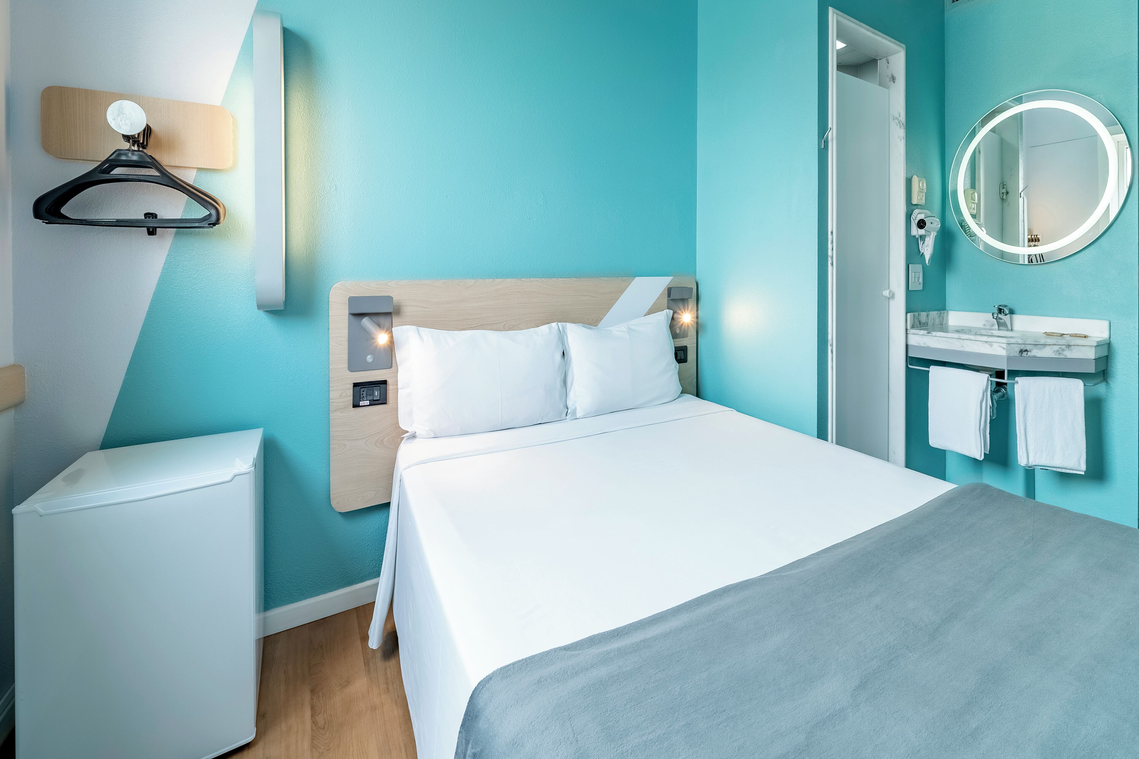 A hotel room with a bed, a small white refrigerator, a wooden headboard, a round mirror, and a bathroom with a marble sink and towel rack.